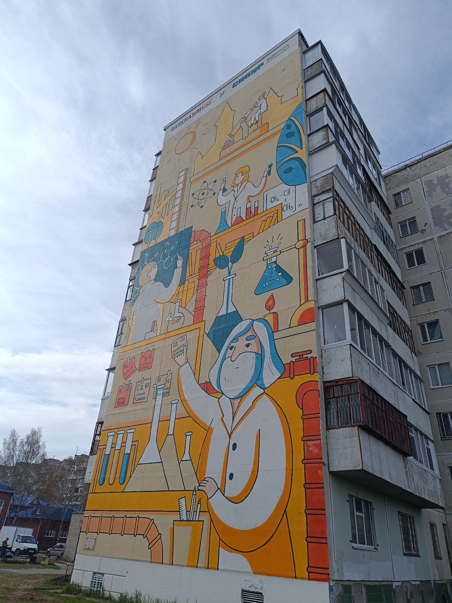 The image captures a tall, multi-story building in Tobolsk, Russia. A large, vibrant mural adorns the building's exterior wall, depicting a series of science-themed illustrations. Prominent figures in lab coats, test tubes, beakers, chemical formulas, and elemental symbols are visible on the mural. The sky is overcast and partly cloudy. The building is surrounded by a patch of grass and some vegetation, with other buildings and vehicles in the background. The mural's design and composition suggest a tribute to scientific exploration and education.