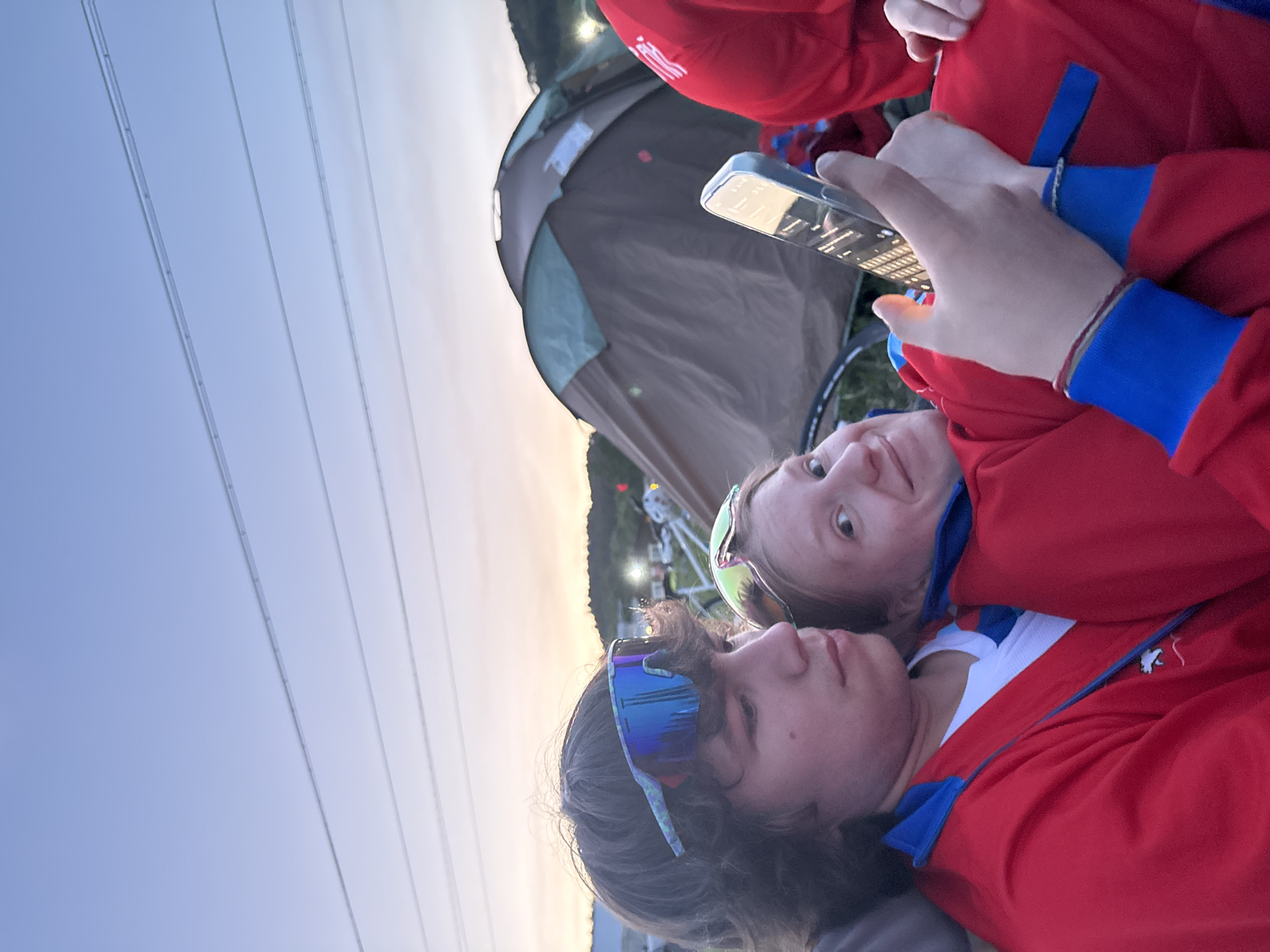 Two individuals are lying down outside, possibly in a camping setting near Khvalynsk, Russia. One person is holding a smartphone, its screen illuminated. A tent is visible in the background. Power lines are stretched across the sky. Both individuals are wearing red shirts with blue accents and sunglasses.