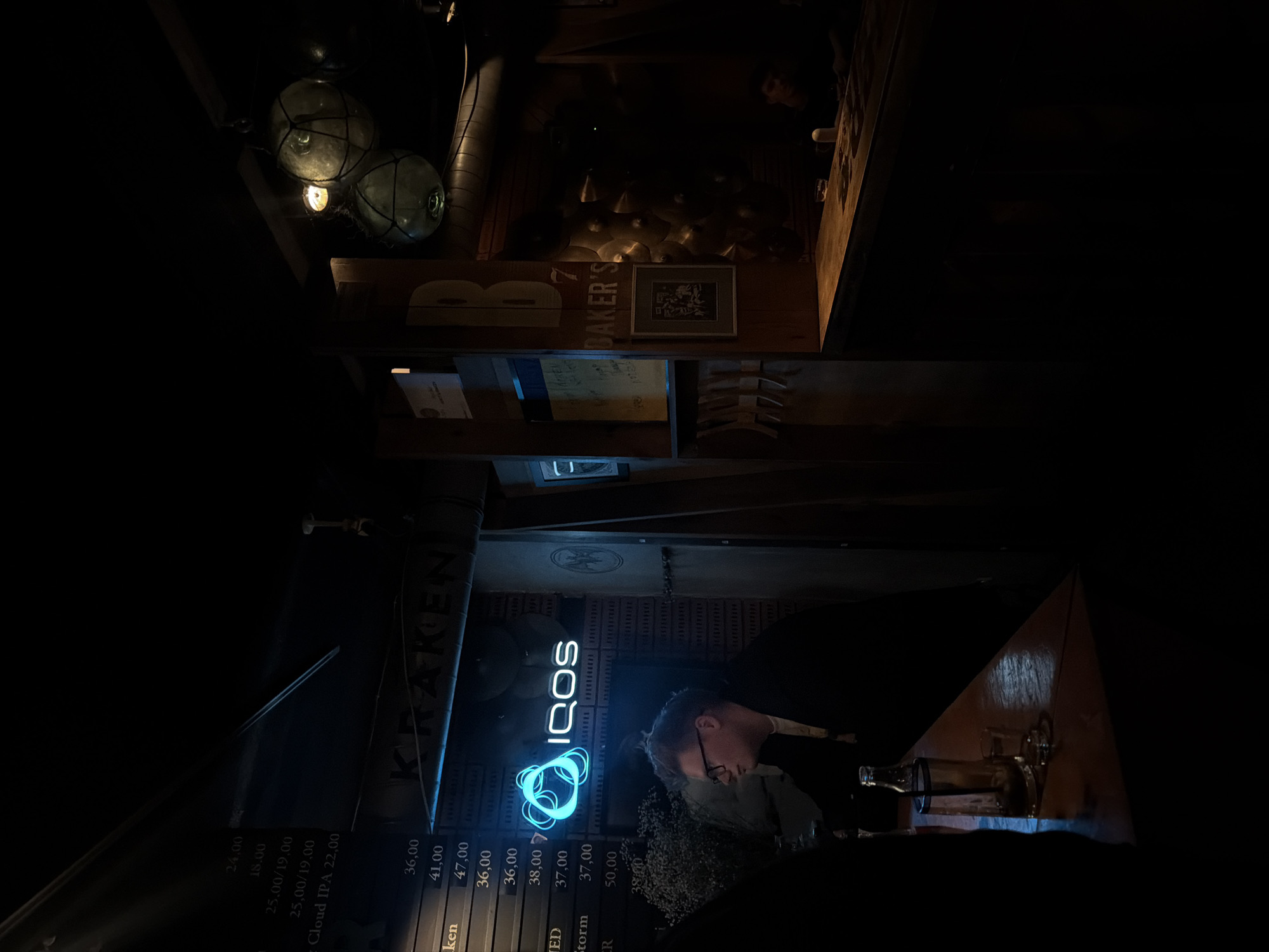 The image shows the interior of a bar or pub. The name "Baker's" is visible on a wooden structure. There are some vintage-style light fixtures hanging from the ceiling. A person is standing near a menu board which advertises "Kraken" and "IQOS" with prices. The interior appears dimly lit, and some decorative items and bottles are visible.
