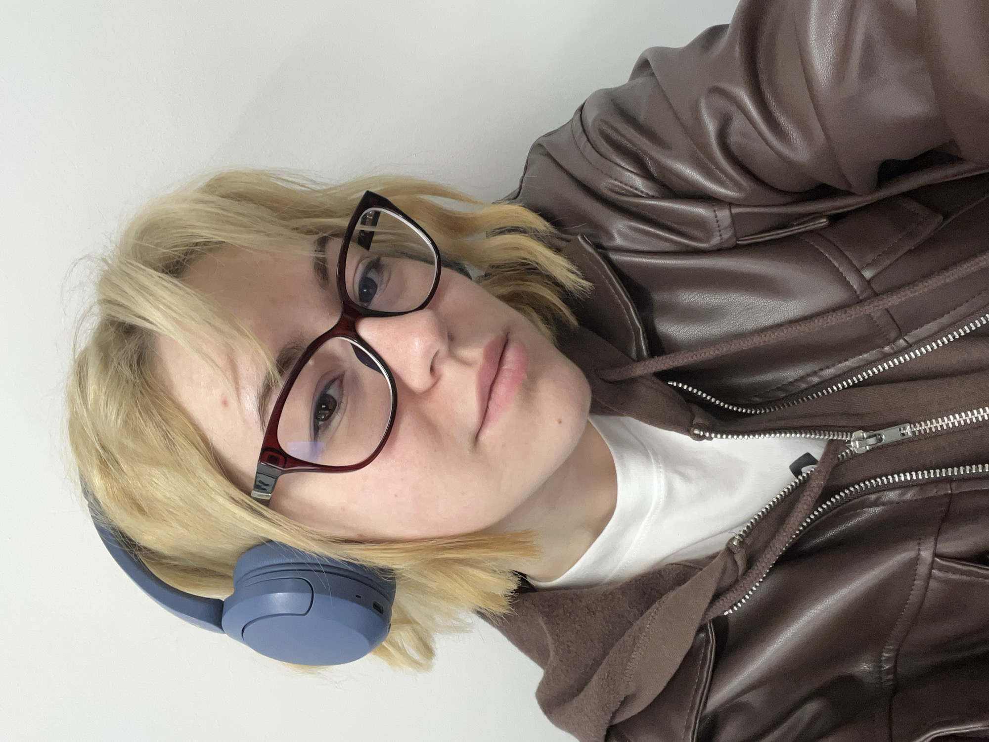 A close-up, eye-level selfie shows a person with light blonde hair. The image is oriented such that the person's head is tilted significantly to the left within the frame. The individual has a fair complexion and a neutral, slightly serious expression, with lips subtly parted. They wear dark-framed glasses with a subtle cat-eye shape, and blue over-ear headphones are positioned around their neck and partially over their left ear.
The person is dressed in layers: a shiny, dark brown faux leather jacket is worn over a brown hooded top with a visible zipper and drawstrings. A plain white t-shirt is visible underneath the hooded top. The background is a plain white wall, suggesting an indoor setting. There are no visible details to confirm the time of day, weather, or the provided location context of Budva, Montenegro. No text is visible in the image.