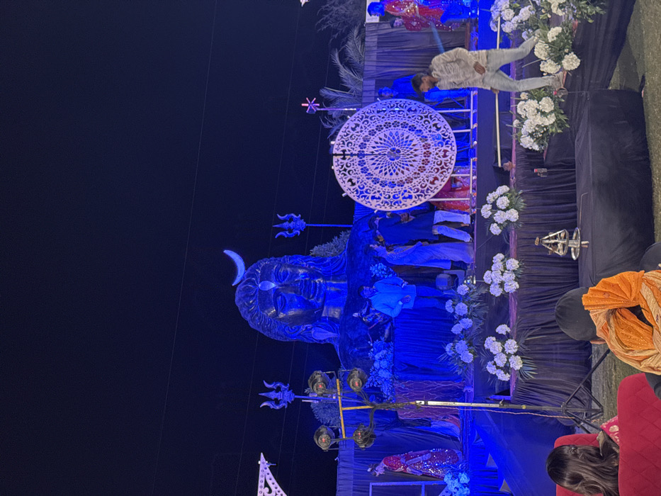 The image displays a sculpture of a deity with a crescent moon head adornment. A large, ornate circular decoration hovers above it. The setup seems to be lit with blue light, and white flower arrangements are placed around the statue. People are gathered around. The photo is oriented vertically.