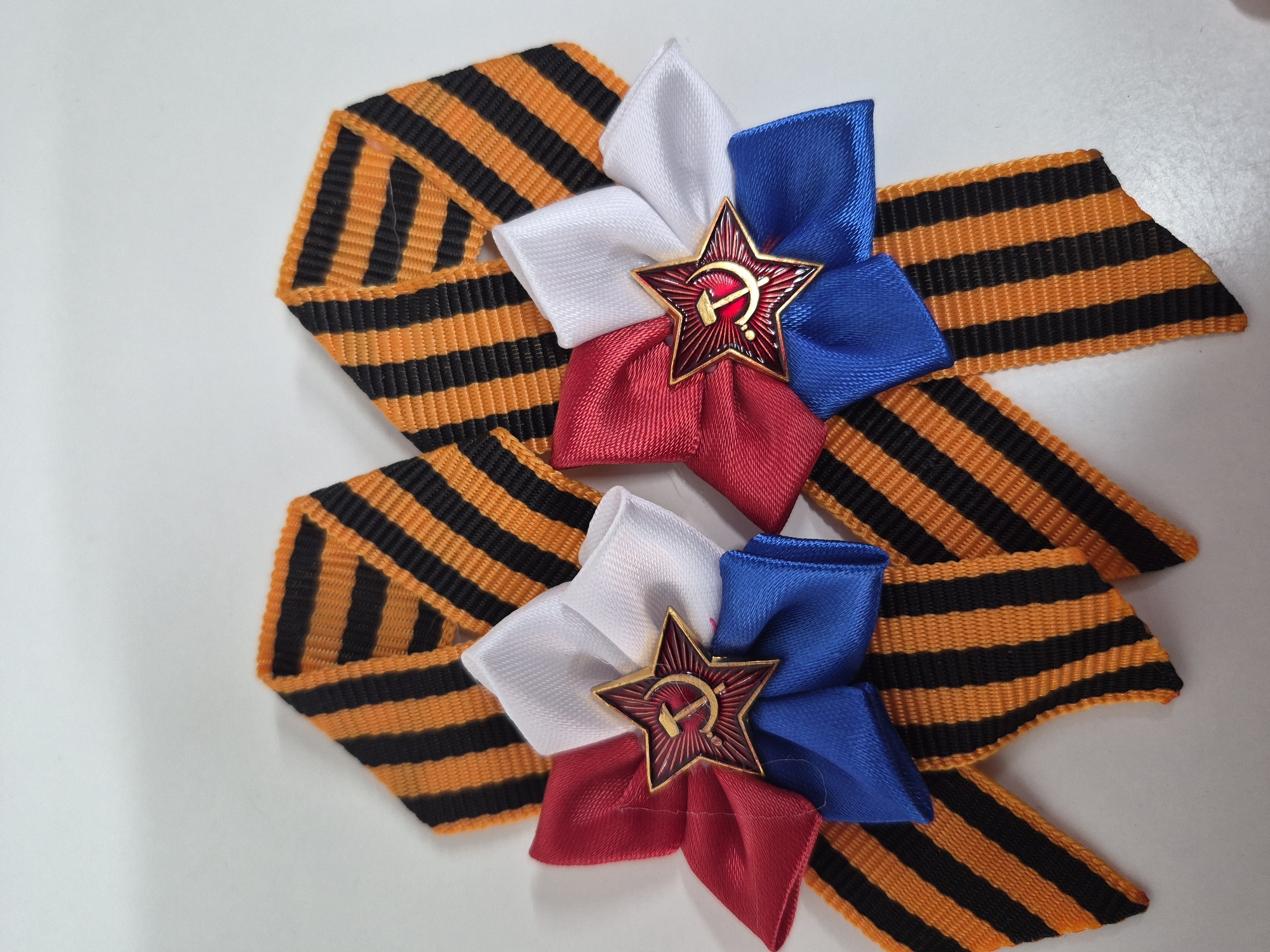 Two orange and black striped ribbons are arranged with decorative rosettes. The rosettes are composed of red, white, and blue ribbons, and each features a red star emblem with a hammer and sickle in the center. The items are displayed on a white surface. There is no indication of Tobolsk, Russia, or any other specific location. The image contains no people, actions, or interactions.