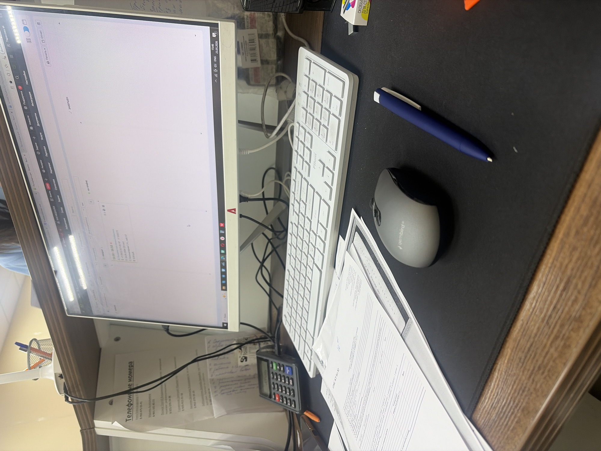 The image shows a workstation setup with a computer monitor displaying a webpage, a white keyboard, a grey wireless mouse, a blue pen, and a calculator. There are also documents stacked on the desk. The monitor shows Russian text, and a sticker indicates "Телефонные номера" ("Telephone numbers").