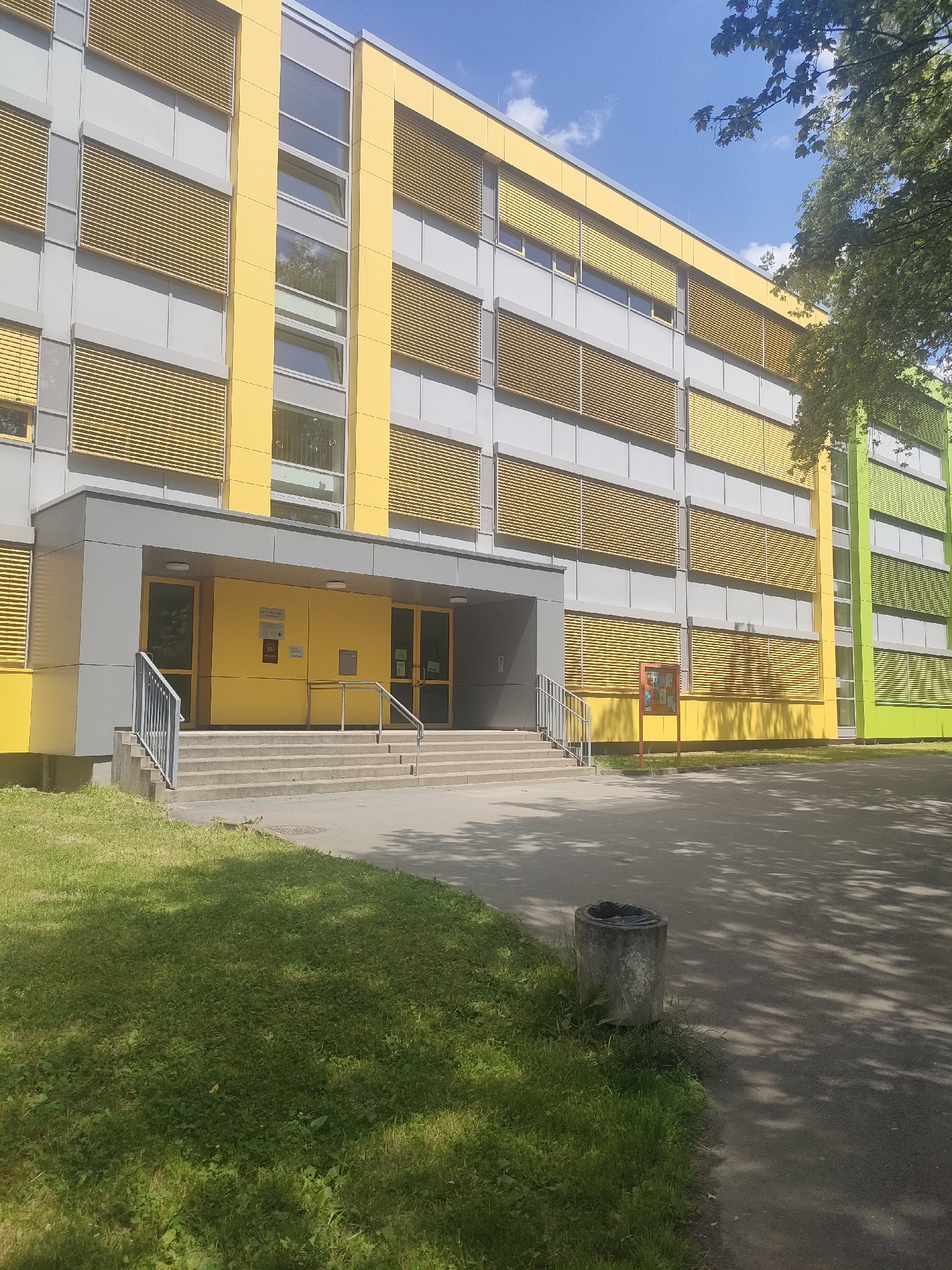 A modern building with a colorful facade stands under a bright blue sky with scattered clouds. The building features alternating bands of gray and vibrant yellow on its upper floors, punctuated by horizontal window blinds that are also yellow. The lower section of the building is a darker gray, leading to a bright yellow entrance area. This entrance is accessed by a wide set of concrete stairs with metal handrails. Two doors, one with glass and another appearing to be a solid yellow door with a mail slot, are visible in the entrance. There are signs on the yellow doors, one appearing to be a white rectangular sign with some text and a red graphic, and another smaller, red sign. A red display board stands to the right of the entrance. To the right of the main building section, the facade transitions to bright green, also with gray sections and yellow blinds. The area in front of the building is a paved walkway and a grassy lawn. A concrete trash receptacle with a black liner sits on the edge of the lawn near the walkway. Sunlight casts dappled shadows on the pavement and grass, suggesting it is a sunny day, likely late morning or early afternoon. The overall scene is clean and orderly, typical of a public or institutional building, possibly a school or office. The location context is Chemnitz, Germany.