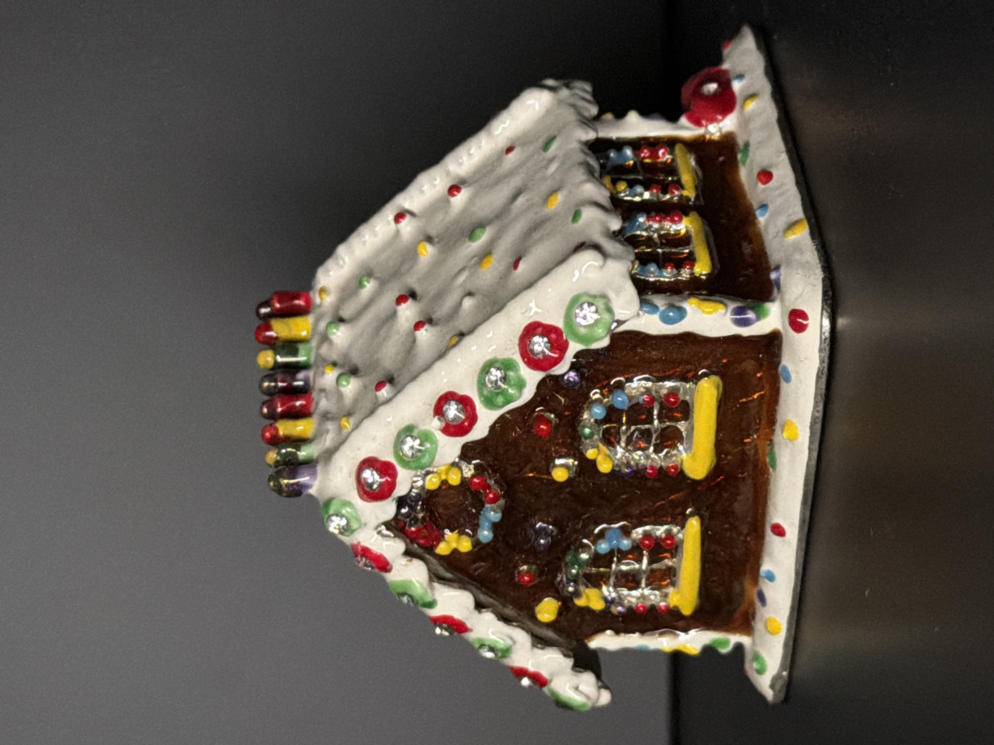 A small, decorative gingerbread house ornament sits on a dark, reflective surface. The ornament is detailed with white icing that mimics snow, red and green candy cane-like trim along the roof edges, and colorful sprinkles scattered across the roof and base. The windows are depicted as panes with yellow sills and red and blue candy accents. A chimney adorned with colorful candle-like figures stands at the peak of the roof. The background is a gradient of dark grey, with the top half being lighter than the bottom half. There are no people present in the image. The location in the city of Baan Bo Phut, Thailand is not discernible from the image.