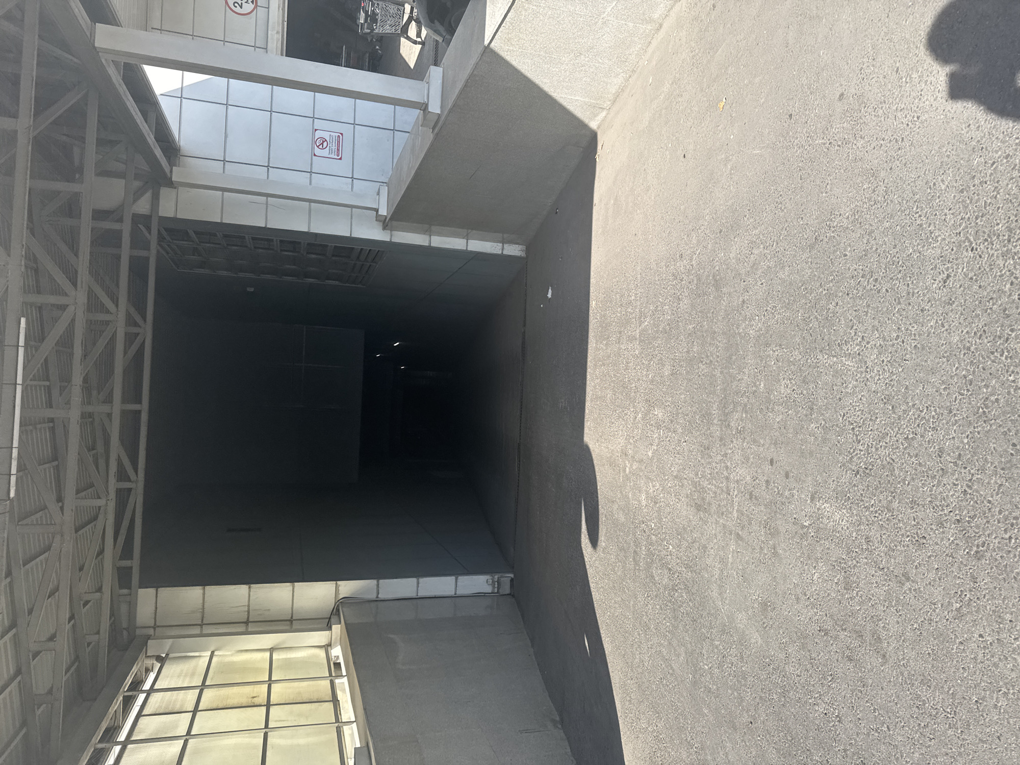 The image shows the entrance to a dark underpass or tunnel. The ground in front of the underpass is paved with asphalt and is partially covered by shadows. The sides of the entrance are lined with concrete walls. To the right of the entrance, there is a white tiled wall with a red and white sign that indicates "No Smoking". Further to the right, there are some shopping carts and a glimpse of vehicles. The overall scene appears to be an outdoor area, possibly a parking garage entrance or a pedestrian underpass, in a city environment. The lighting suggests it is daytime, with strong sunlight casting sharp shadows.