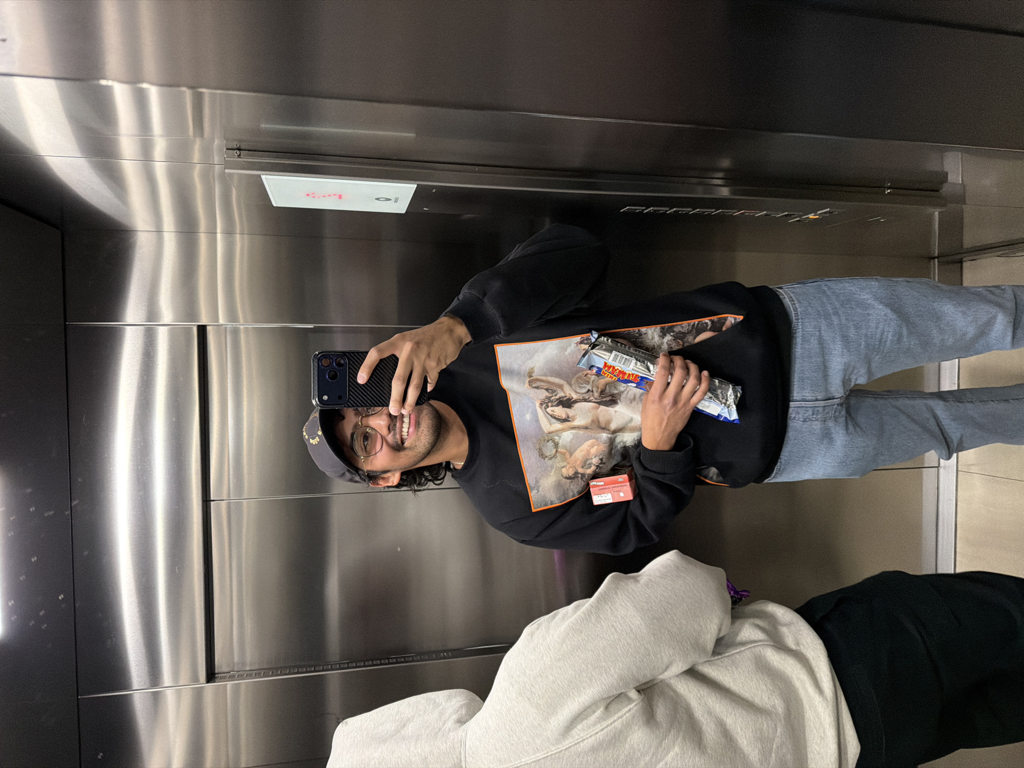 A person takes a selfie in an elevator. The person is a young adult male with dark hair and a beard, wearing glasses, a cap, a black sweatshirt with a Renaissance painting on the front, light blue jeans, and white sneakers. In his hands, he holds a candy bar. The elevator has stainless steel walls. Behind the person is another individual with light grey hoodie and black pants. The location is San Andrés Cholula, Mexico.