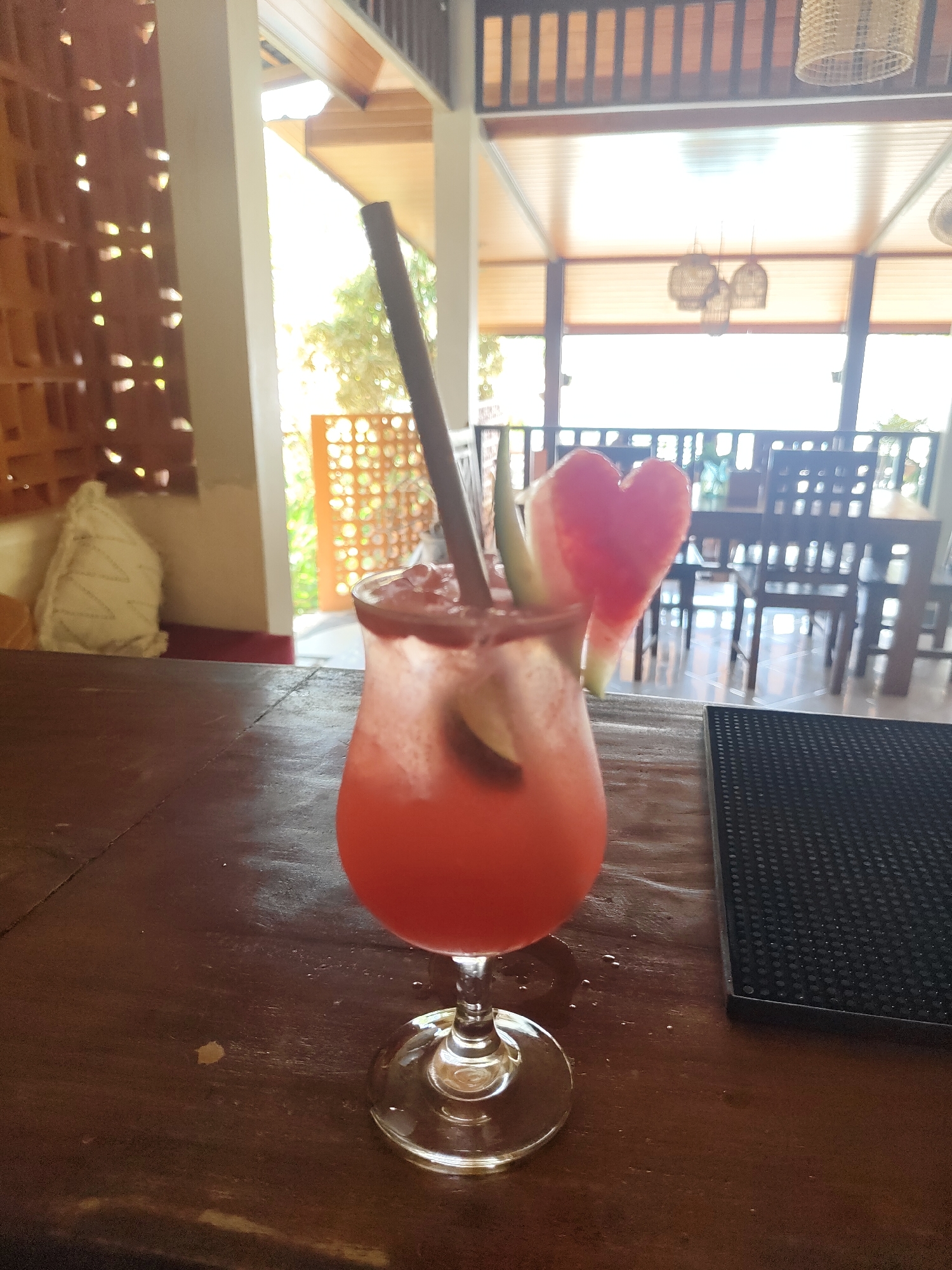 A vibrant, reddish-pink cocktail in a stemmed hurricane glass is the central focus, positioned slightly to the left in the foreground. The drink is garnished with a dark straw and a distinctive heart-shaped slice of watermelon, along with what appears to be a smaller piece of fruit or rind inside the glass. The glass rests on a dark, polished wooden bar or table, which shows some subtle grain and minor imperfections. To the right of the cocktail, a textured black rubber bar mat is visible.
The setting is an airy, semi-open restaurant or bar, characterized by natural light flooding in from the background. The architectural style features wooden elements, including a slatted railing across the top and in the mid-ground, and wooden beams on the ceiling. Decorative woven light fixtures hang from the ceiling. To the left, a wall with an intricate pattern of square cut-outs allows light to filter through, and a white, textured cushion is visible on a seating area. Beyond the main space, bright sunlight illuminates an outdoor area, with hints of greenery and further patterned walls. Dark wooden tables and chairs are arranged in the background, suggesting a dining or lounge area.
The scene depicts a peaceful, daytime ambiance, likely in a tropical or resort location like Kuta Selatan, Indonesia, given the open design and light-filled environment. There are no people visible. The bright light suggests it is sunny outside. No discernible text is present in the image.