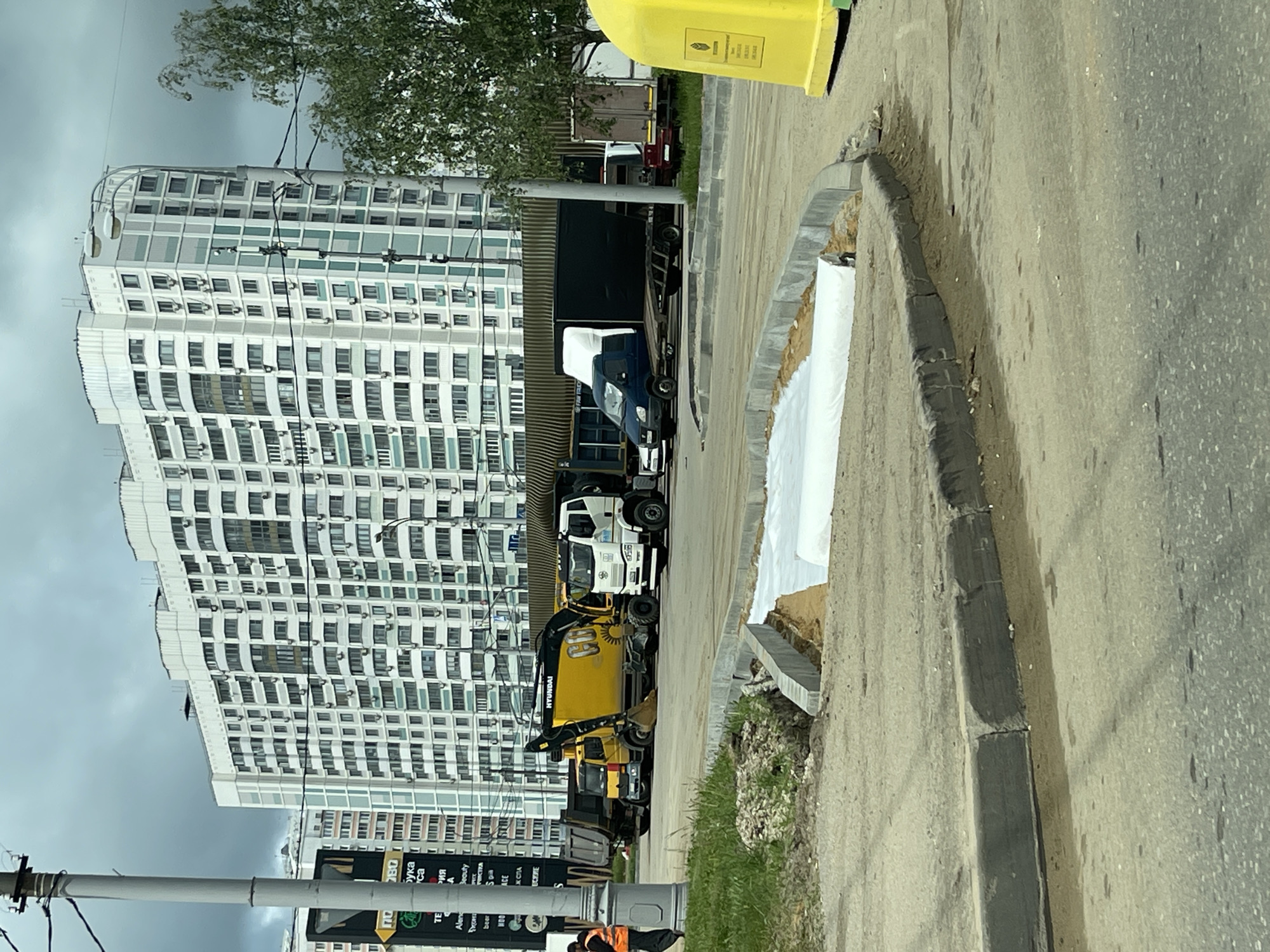 The image shows a street scene in Moscow, Russia, dominated by a large, modern, high-rise apartment building with numerous windows. A street runs parallel to the building's base, and along the street's edge, various construction and utility vehicles are parked. These include a yellow excavator with the brand name "Hyundai," several flatbed trucks, and a smaller blue truck covered with a white tarp. A yellow trash bin is visible on the sidewalk. Roadside work is underway, as indicated by the exposed earth and construction materials. The sky is overcast, suggesting a cloudy day. In the foreground, a utility pole and street signage can be seen, indicating the urban nature of the location.