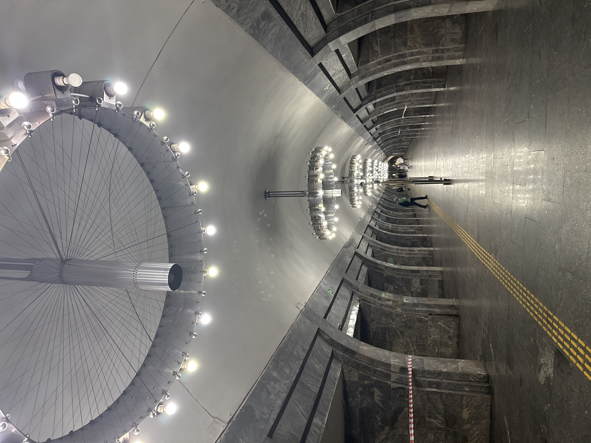 The image shows the interior of a Kyiv Metro station, likely a deep underground station given the architecture. The station features a long, vaulted tunnel with arched alcoves along the sides, clad in dark, patterned stone. The ceiling is high and covered in a light-colored material, from which hang elaborate, circular chandeliers with numerous exposed light bulbs. These chandeliers are a prominent feature, with at least two visible in the distance, reflecting on the polished floor. A raised yellow tactile paving strip runs along the edge of the platform. In the foreground, red and white caution tape is strung across one of the alcoves, indicating a restricted area. Several people are visible in the distance, walking along the platform or waiting, but they are small and indistinct. The lighting from the chandeliers creates a somewhat dramatic and cavernous atmosphere.