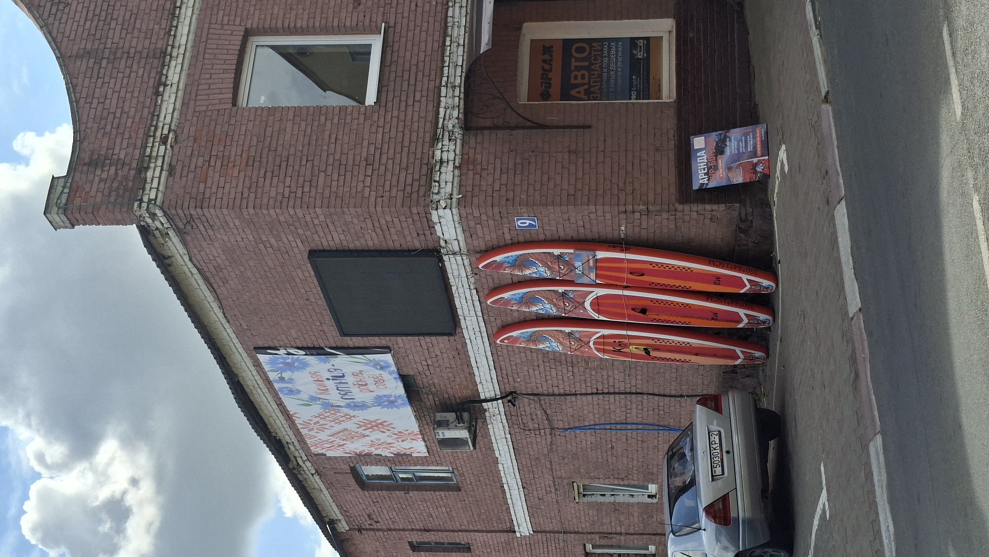 The image depicts the exterior of a brick building located in the city of Pastavy, Belarus. Three orange paddleboards with black and white graphics are propped upright against the wall of the building. To the left of the paddleboards, a silver car is parked on a paved surface. Above the paddleboards, on the brick wall, is a blue and white banner with floral patterns and Cyrillic text. A black electronic display board is mounted higher on the wall, to the left of a window. A sign with Cyrillic text and an image is placed on the ground near the paddleboards. The building has multiple windows and an air conditioning unit mounted on the wall. The sky above is partly cloudy.