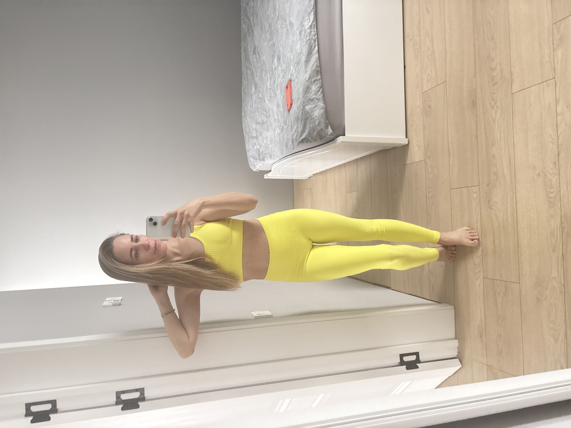 A woman with long blonde hair stands in a room, facing the camera. She is wearing a matching yellow athletic set. She holds a smartphone in her hand and appears to be taking a selfie in front of a mirror. Behind her, the room contains a bed with gray bedding and a wooden floor. The location is not discernible.