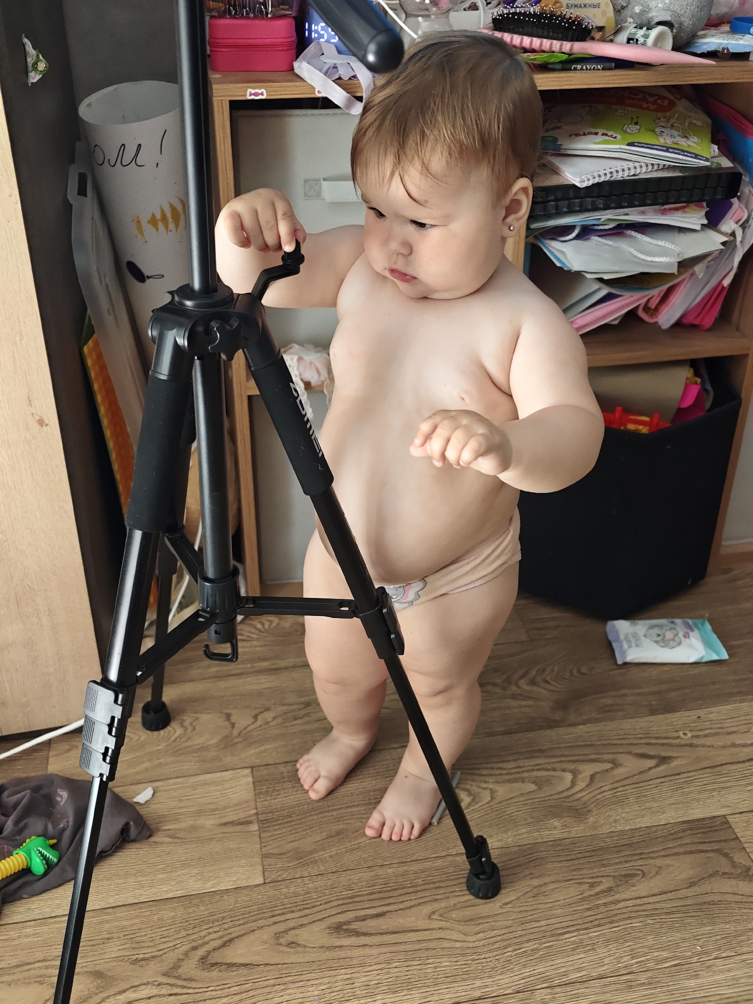 A young child, likely a toddler, stands in a room with wooden flooring. The child is unclothed except for a diaper and is facing a black tripod. The child is interacting with a lever on the tripod, suggesting they are exploring or playing with it. The background reveals a shelf unit filled with books, papers, and various other items, indicating a home environment. A toy dinosaur is visible on the floor to the left of the tripod. On the floor to the right of the child, a package of baby wipes can be seen. The overall impression is a candid snapshot of a child's curious exploration within a cluttered living space. The time of day is not discernible, and no weather cues are present. There is text visible on a roll of paper in the background, which appears to be in Russian, reading "боль!" (bol', meaning "pain!").