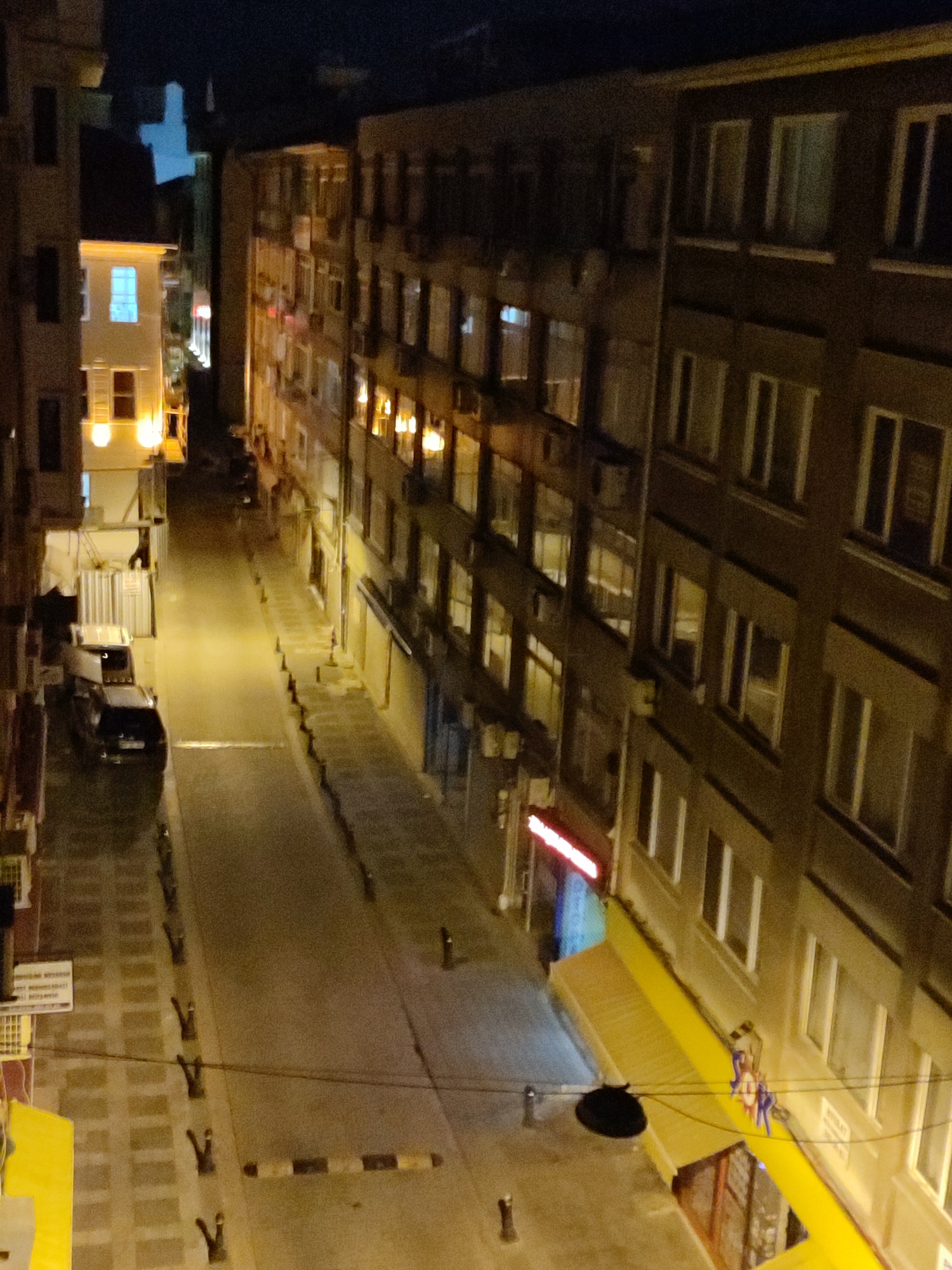 The image shows a dimly lit street scene at night in Kadıköy, Turkey. Several multi-story buildings line both sides of a narrow street. The buildings have numerous windows, many of which are illuminated, indicating occupied apartments or businesses. One building on the left has a bright yellow awning extending over a storefront.
On the street, there are a few parked cars and traffic cones, suggesting a low-traffic area. In the distance, a neon sign glows red. Several small, dark figures, possibly people, are visible on the street, some standing and some appearing to be walking. There is also a black, circular object hanging from wires, which could be a streetlamp. The overall impression is of a quiet urban environment at night.