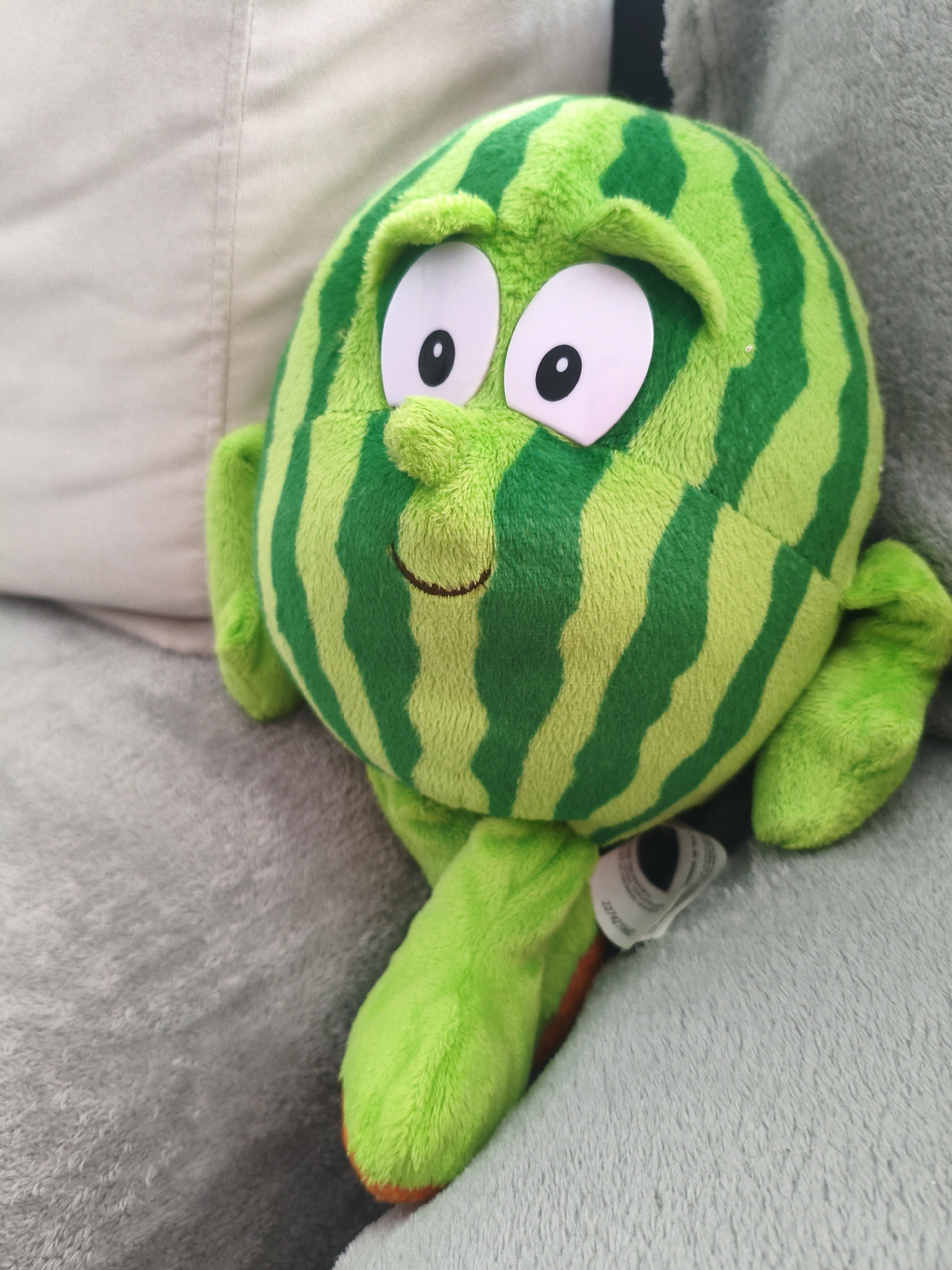 A plush toy resembling a green watermelon with darker green stripes, large white eyes with black pupils, a small nose, and a stitched-on smile sits on a gray, textured surface, likely a couch. The toy has small arms and a leg extended forward. Part of a white pillow is visible in the background. A white tag is attached to the toy. The image provides no information to indicate a location in Plovdiv, Bulgaria.
