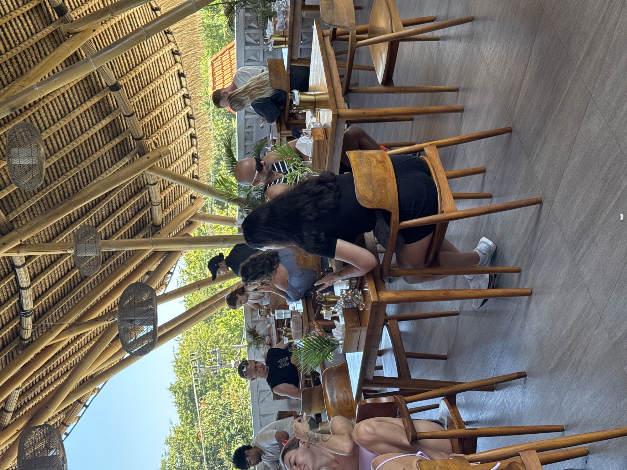 The image captures a group of people dining at a restaurant in Kuta Selatan, Indonesia. The structure of the restaurant is constructed from bamboo, forming a peaked roof with hanging fans. Several individuals are seated at wooden tables, engaged in conversation and interacting with their meals. There are individuals sitting on the chairs, and the setting appears to be a relaxed, open-air dining area. The outdoor environment is visible in the background, with lush greenery and a clear sky. The scene suggests a casual dining atmosphere.