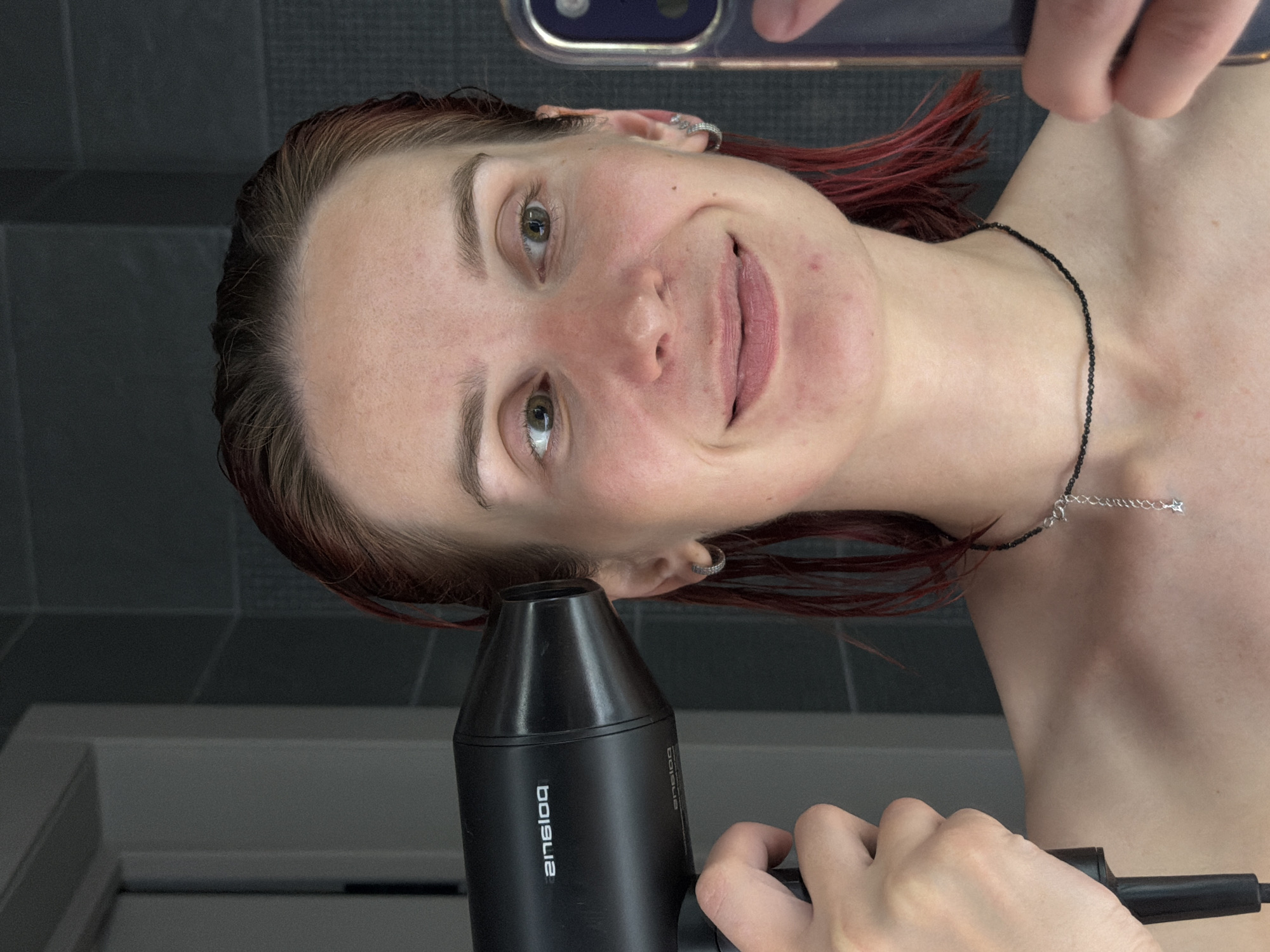 A young woman is holding a black hair dryer to her head while using a smartphone to take a selfie in a bathroom. The woman has fair skin, light brown eyebrows, and green eyes. Her hair is damp and reddish-brown, with some strands dyed red. She is wearing a black beaded necklace with a small silver charm. The bathroom has gray tiled walls and a white doorframe is partially visible. The hair dryer has the word "belsiq" printed on it. The location is unknown.