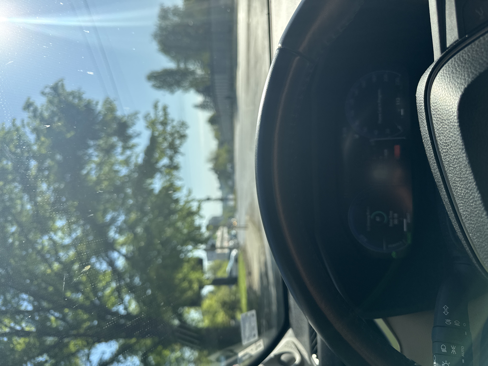 A photo taken from inside a car, looking out the windshield. The road is lined with green trees under a blue sky, the sun is shining. The car's steering wheel is in the right side of the image. The dashboard and turn signal are visible. The location is not discernible from the image.