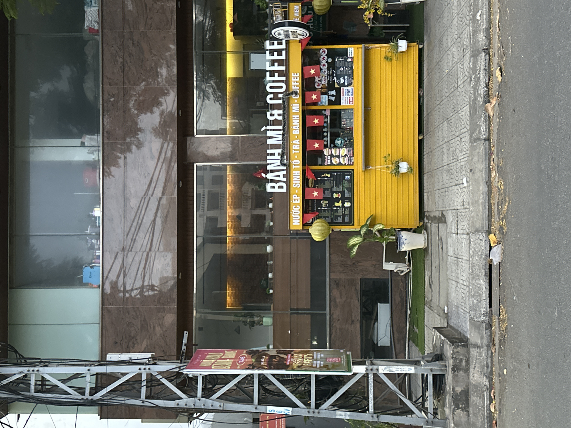 An outdoor stall with a bright yellow facade, labeled "BÁNH MÌ & COFFEE" in large white lettering, stands on a sidewalk in Da Nang, Vietnam. The stall displays menus with images of food and drinks, and is adorned with small red flags featuring yellow stars. A circular sign on the right side of the stall also reads "BÁNH MÌ CA PHÊ". Two potted plants hang from the front of the stall, and a larger potted plant stands to the right. The stall is situated in front of a modern building with large glass windows and a brown marble exterior. A utility pole with numerous wires stands to the left of the stall. The street in front of the sidewalk is paved and appears to be a road. The scene is captured from a street-level perspective, looking slightly upwards towards the stall and building.