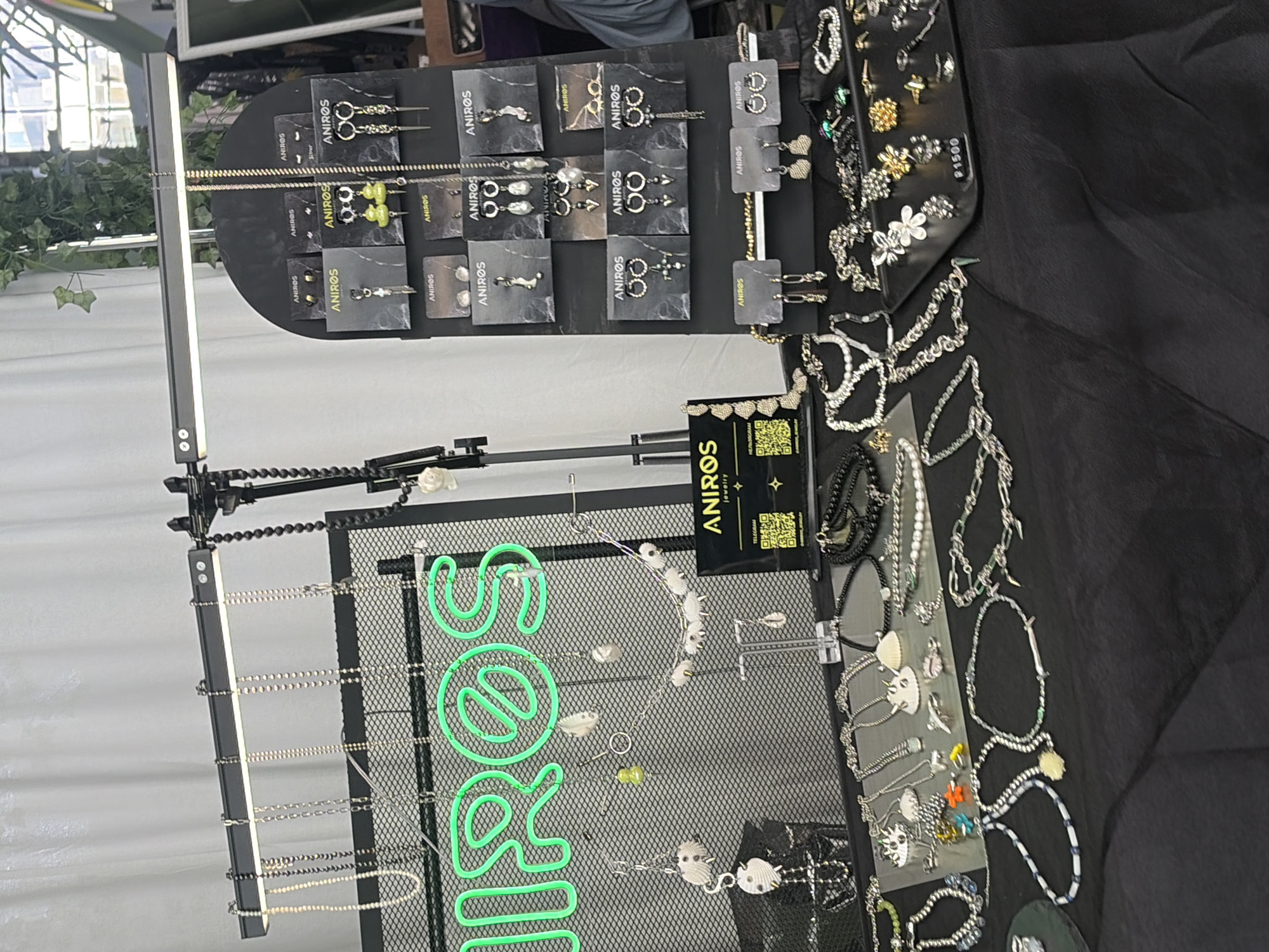 This detailed image captures an indoor jewelry stall, likely at a market or fair, featuring a wide assortment of necklaces, earrings, and bracelets. The display is set up on a table draped with a black fabric covering, and overhead, a horizontal black light bar provides illumination.
In the midground, a black metal mesh panel acts as a primary display surface, featuring a prominent neon green sign spelling "ANIROS" with a stylized diagonal line through the 'O'. Several necklaces, many with white shell or pearl pendants, are suspended from the top bar of this panel and directly from the mesh. To the right, a tall, arched black display stand showcases numerous pairs of earrings, each presented on small black cards branded with "ANIROS". The earrings exhibit diverse styles, including various hoops, studs, and dangling designs, incorporating elements such as pearls, green-colored beads, and metallic charms.
The table in the foreground is densely covered with additional jewelry items. These include an array of silver-toned chain necklaces, beaded bracelets, and further pieces adorned with natural elements like shells. On the right side of the table, a smaller black tray holds a collection of rings and earrings, with one item bearing a white price tag displaying "P1500". QR codes are visible on small cards near the display, associated with "ANIROS jewelry" and mentioning "Telegram".
The background consists of a plain white fabric drape, possibly a partition or backdrop. In the upper right corner, some green foliage, indicative of a decorative plant, is visible. Further to the right, a multi-paned window with dark, industrial-style framing suggests the venue's architecture. Based on the branding "ANIROS jewelry" and common market locations for similar vendors, the scene is situated in the city of Saint Petersburg, Russia, likely within a venue known for artisan markets, such as Sevkabel Port. No people are visible within the frame.
