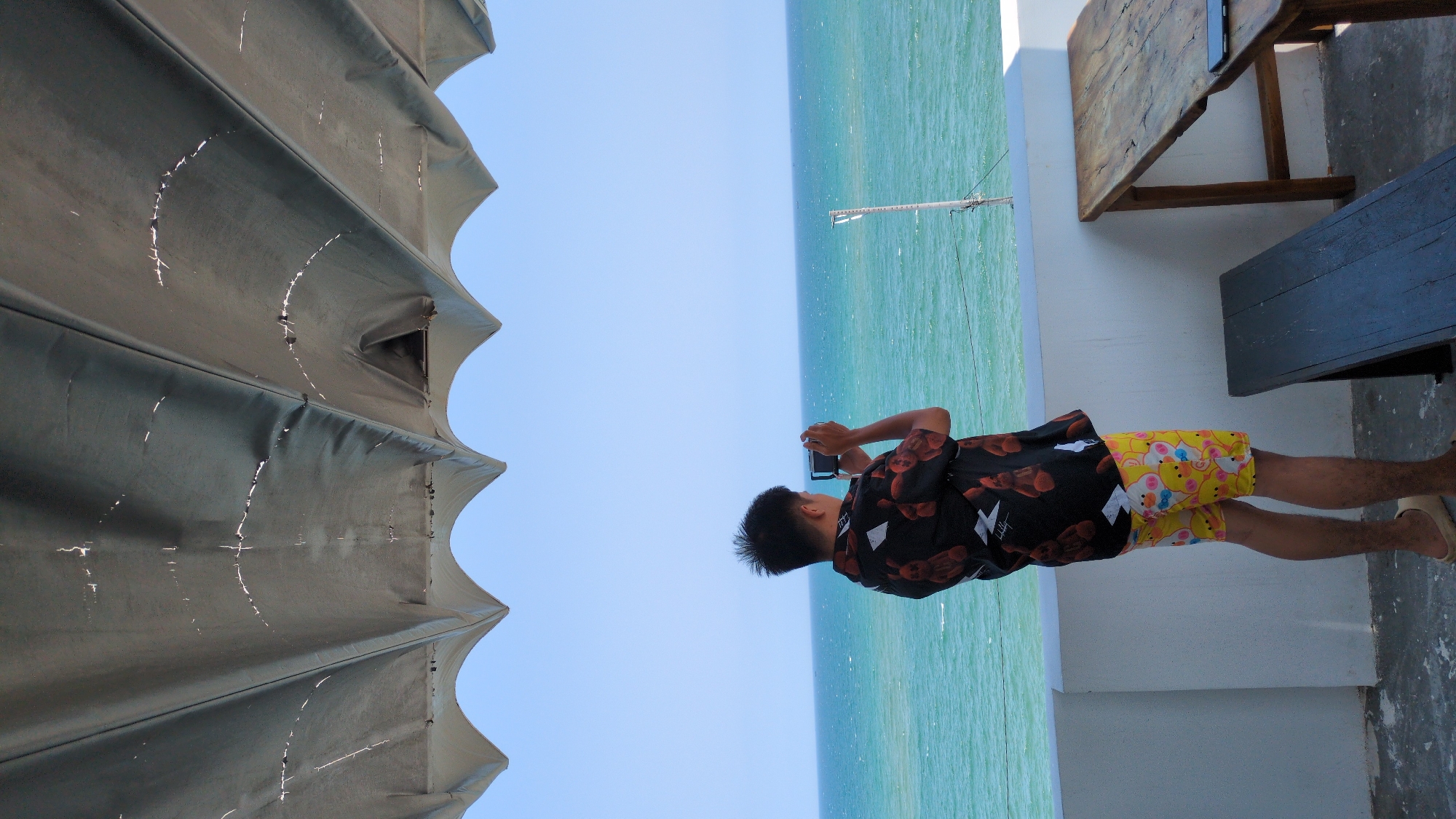 A man stands by the sea in Phan Rang – Tháp Chàm, Vietnam, taking a photo with his smartphone. He is wearing a black shirt with a red rose pattern and yellow shorts with a cartoon pattern. He is also wearing sandals. The sea is turquoise, and the sky is clear blue. To the left, there is a tent-like structure, and to the right, there is a wooden table and a bench. The ground is made of concrete.