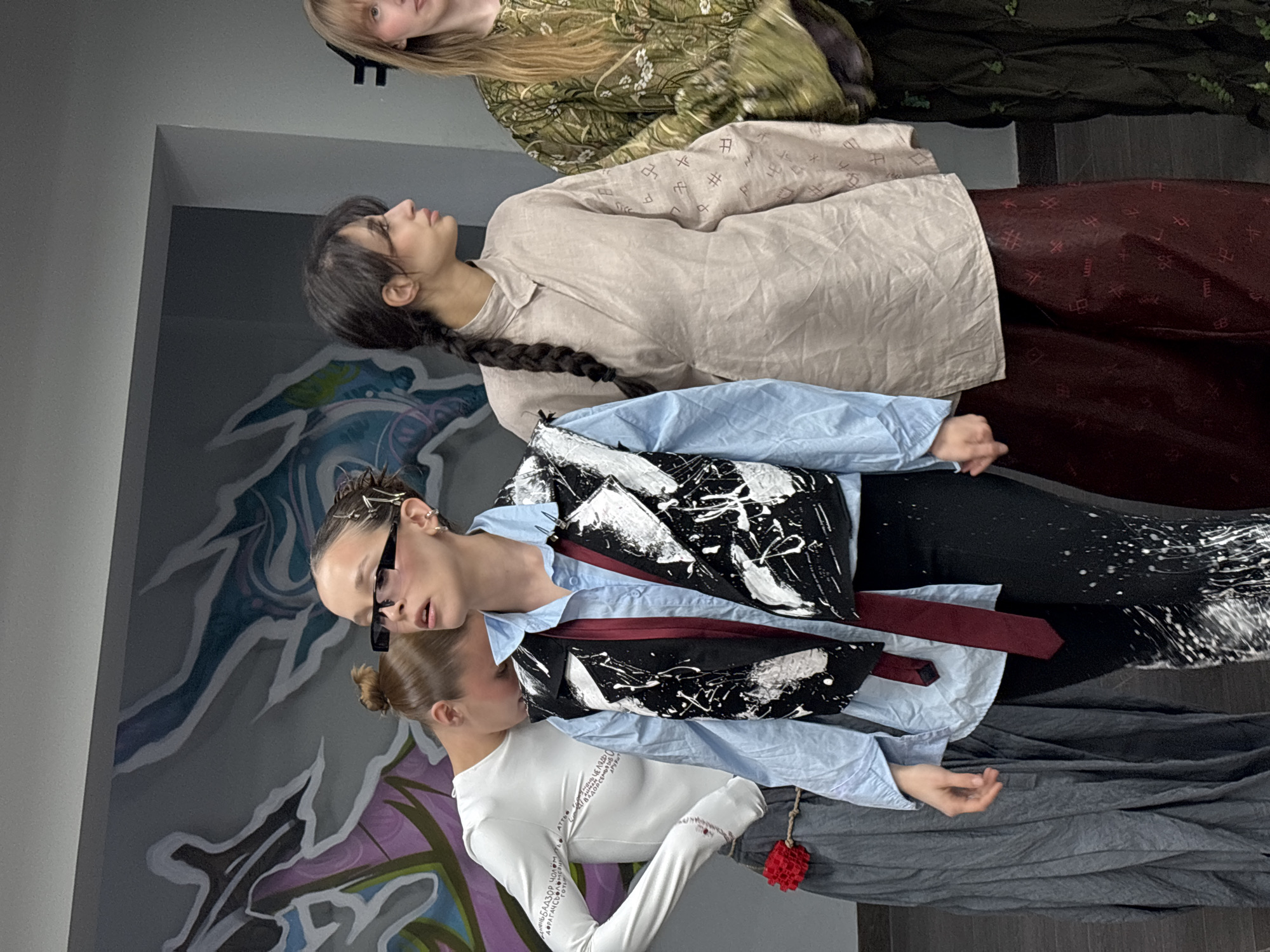 The image captures a fashion-forward scene indoors, likely a photoshoot. Three women and a girl are featured, each in a distinct outfit with varying styles. The first woman wears a flowing green dress with subtle floral patterns. The second woman is dressed in a beige shirt with unique markings and a dark red skirt. The third woman is wearing a light blue shirt with a black and white vest and a red tie. The girl wears a long-sleeved white shirt, grey pants, and is holding a small red accessory. The background includes a wall with graffiti-style artwork. In the lower left corner of the image, there is a jumble of text including "Perm, Russia".