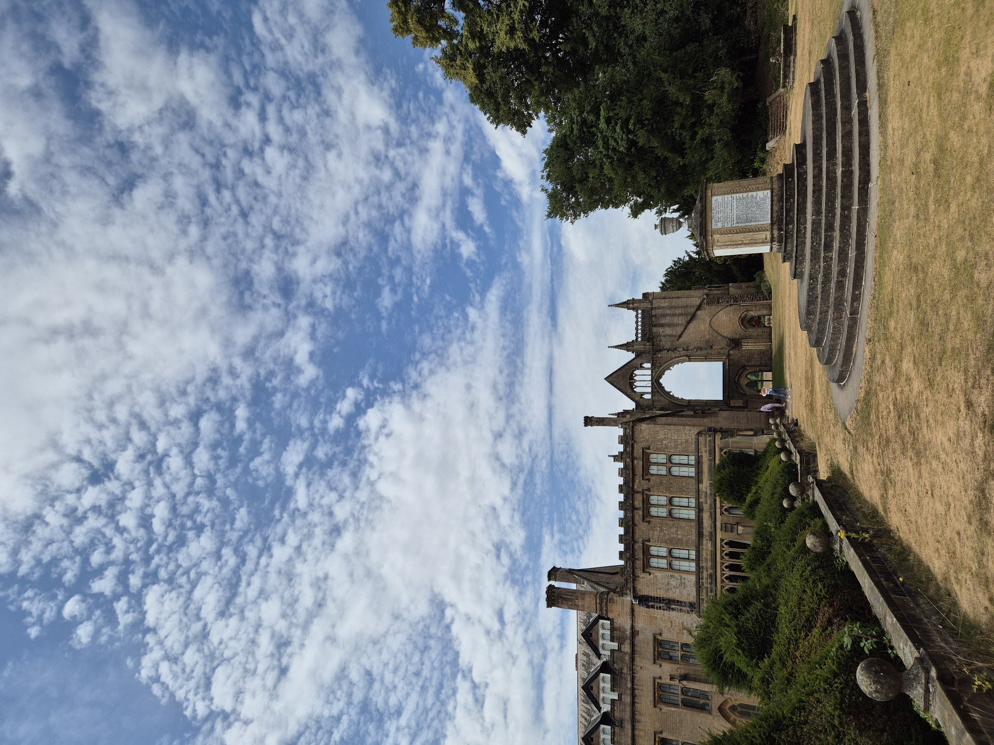 The image depicts a wide-angle view of the ruins of Newstead Abbey in Gedling, United Kingdom. The sky above is a vibrant blue, filled with scattered, fluffy white clouds. In the foreground, a circular, tiered stone structure anchors the scene, topped with a stone monument bearing inscriptions. The ground surrounding this structure is covered in dry, yellow grass, indicating a summer season.
To the left of the monument, a section of the abbey's main building is visible. This part of the structure features a stone facade with multiple windows and ornate architectural details. Adjacent to this building, a neatly trimmed, dense green hedge creates a stark contrast with the dry grass.
In the center of the image, the iconic arched window of the abbey ruins stands prominently. Two individuals are visible near this archway, appearing to be casually observing the surroundings. To the right of the ruins, a large, mature tree with lush green foliage dominates the background, with its branches reaching towards the sky. Stone steps are also visible leading up to a higher level of the grounds behind the tree. The overall impression is one of historical grandeur set within a natural landscape under a bright, cloudy sky.
