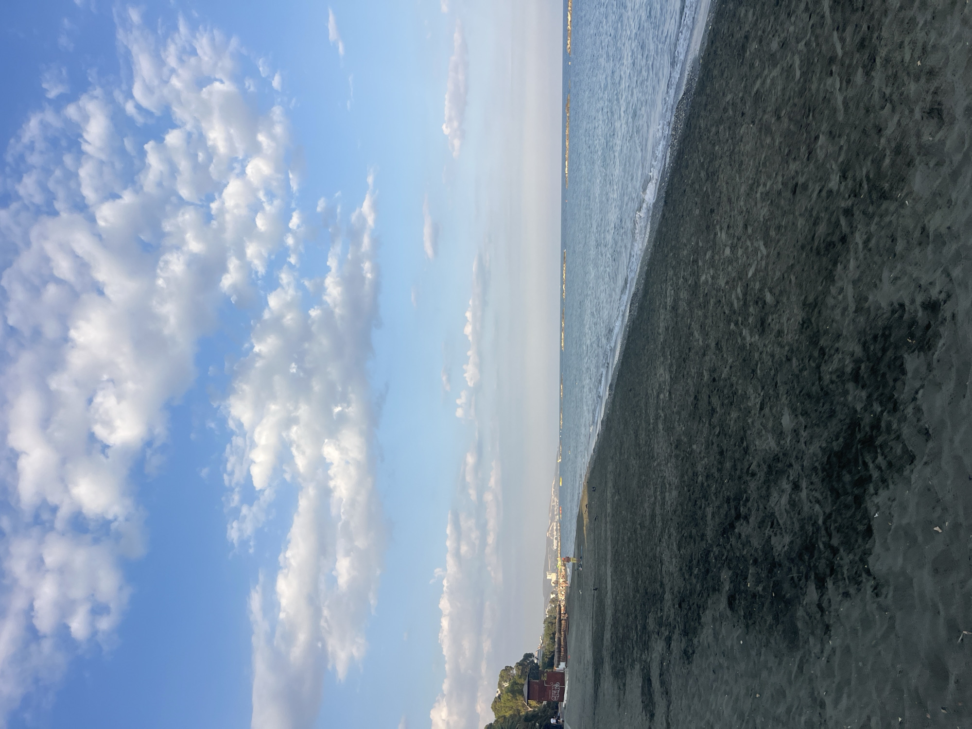 The image shows a beach scene in Agios Tychona Community, Cyprus, under a bright blue sky with scattered white clouds. The foreground is dominated by dark, wet sand, indicating the edge of the receding tide. The sea is calm, with gentle waves lapping at the shore. In the midground, there are several lines of offshore breakwaters, marked by clusters of rocks. Along the coastline in the distance, there are buildings, trees, and what appears to be a small beachside establishment with a red roof. A few people are visible on the beach, scattered and engaged in various activities, including walking. The overall impression is a serene coastal environment.