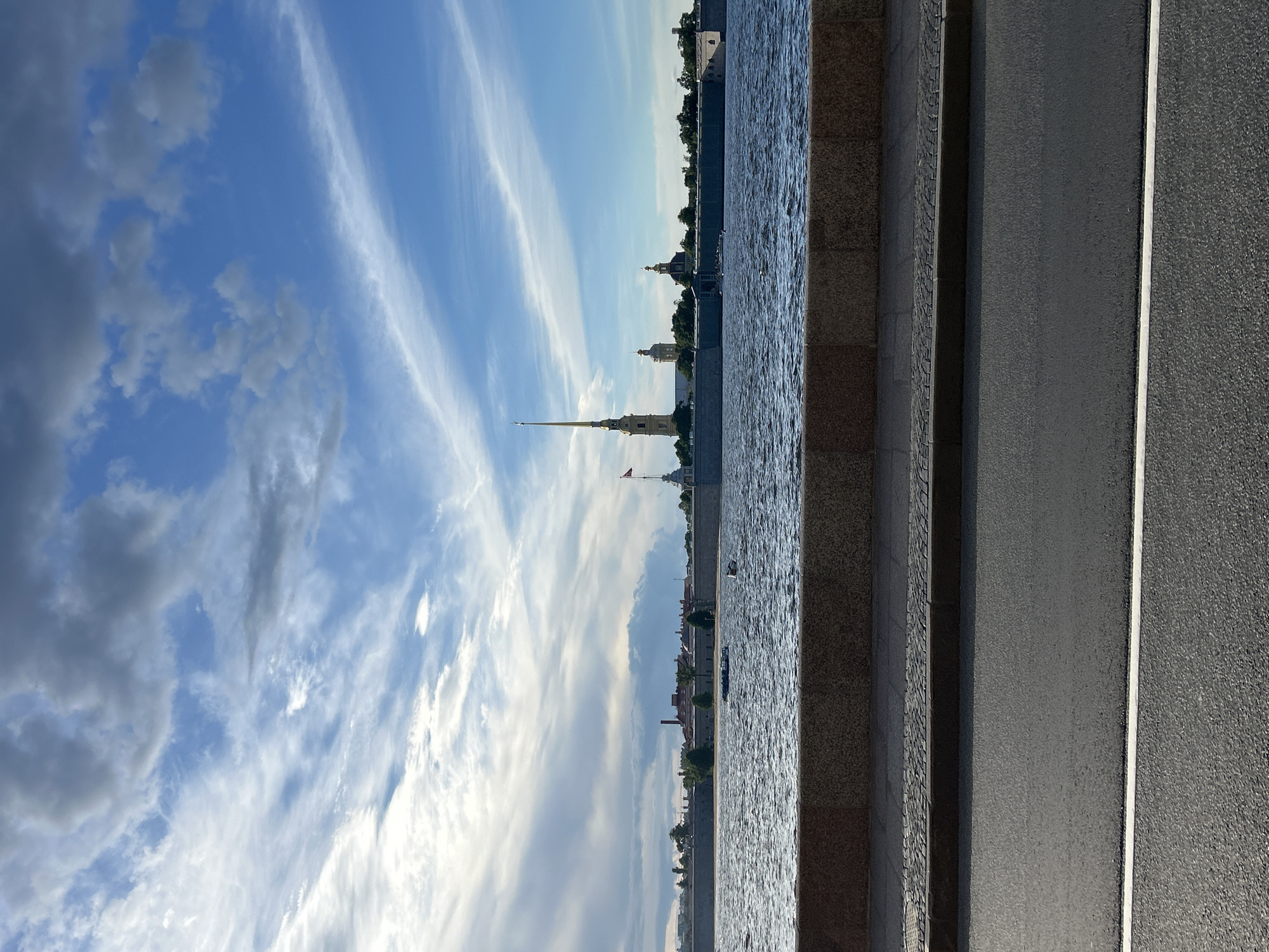 This media file is a landscape orientation photograph depicting a city skyline across a body of water. The sky is a vibrant blue with wispy white clouds, suggesting fair weather. The sun appears to be low in the sky, casting a warm light, indicating either early morning or late afternoon.
The scene is divided into several distinct horizontal bands. The top band is dominated by the expansive sky. Below this is the shimmering surface of the water, which reflects the sky and the buildings along the opposite bank. The buildings in the distance are varied, with several prominent domed structures and a tall, slender spire topped with a flag. These architectural elements suggest a historic European city. A line of trees is visible along the waterfront. A few small boats are present on the water.
In the foreground, there is a paved walkway or embankment, constructed with large granite blocks and a textured stone surface. To the right of this embankment is a dark asphalt road, marked with a thin white line. The contrast between the textured embankment and the smooth road is noticeable.
There are no people or animals visible in the frame. The overall mood is peaceful and serene, capturing a moment of quiet beauty in an urban environment. The visual cues suggest a location with a rich history and significant landmarks, likely a waterfront in a well-established city. No text is visible in the image.