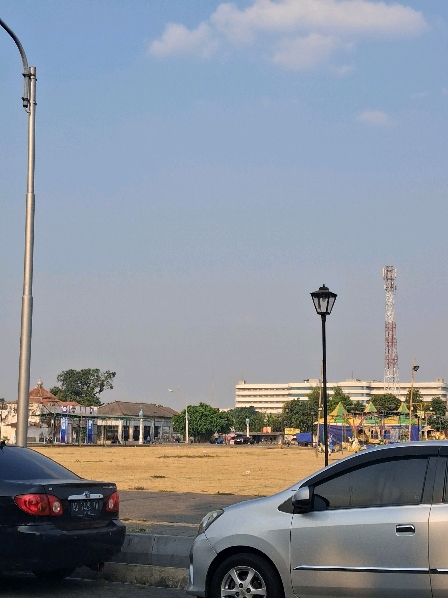 The image depicts an outdoor scene, likely in the city of Surakarta, Indonesia, given the context provided. In the foreground, two cars are parked on a paved surface with a concrete barrier. The car on the left is black, and its license plate reads "AD 1429 TH". The car on the right is silver.
Behind the cars, a large, dry, open field stretches into the middle ground. Several buildings are visible on the far side of the field. To the left, there is a low-rise building with traditional architectural elements, including a domed roof. A tall, silver lamppost stands to the left of the field, a prominent feature in the composition. In the center of the frame, a black lamppost with a decorative top is visible, and further to the right, a tall, red and white telecommunications tower dominates the skyline. Behind the field and the buildings, a large, multi-story white building with many windows is visible. A small, colorful amusement ride area with tents is also present.
The sky is clear with a few scattered clouds. The lighting suggests it is either morning or late afternoon. There are a few people visible in the distance near the buildings and the amusement ride, but their actions are not discernible. The overall scene portrays a public space in an urban environment, possibly a park or a town square, adjacent to a field.