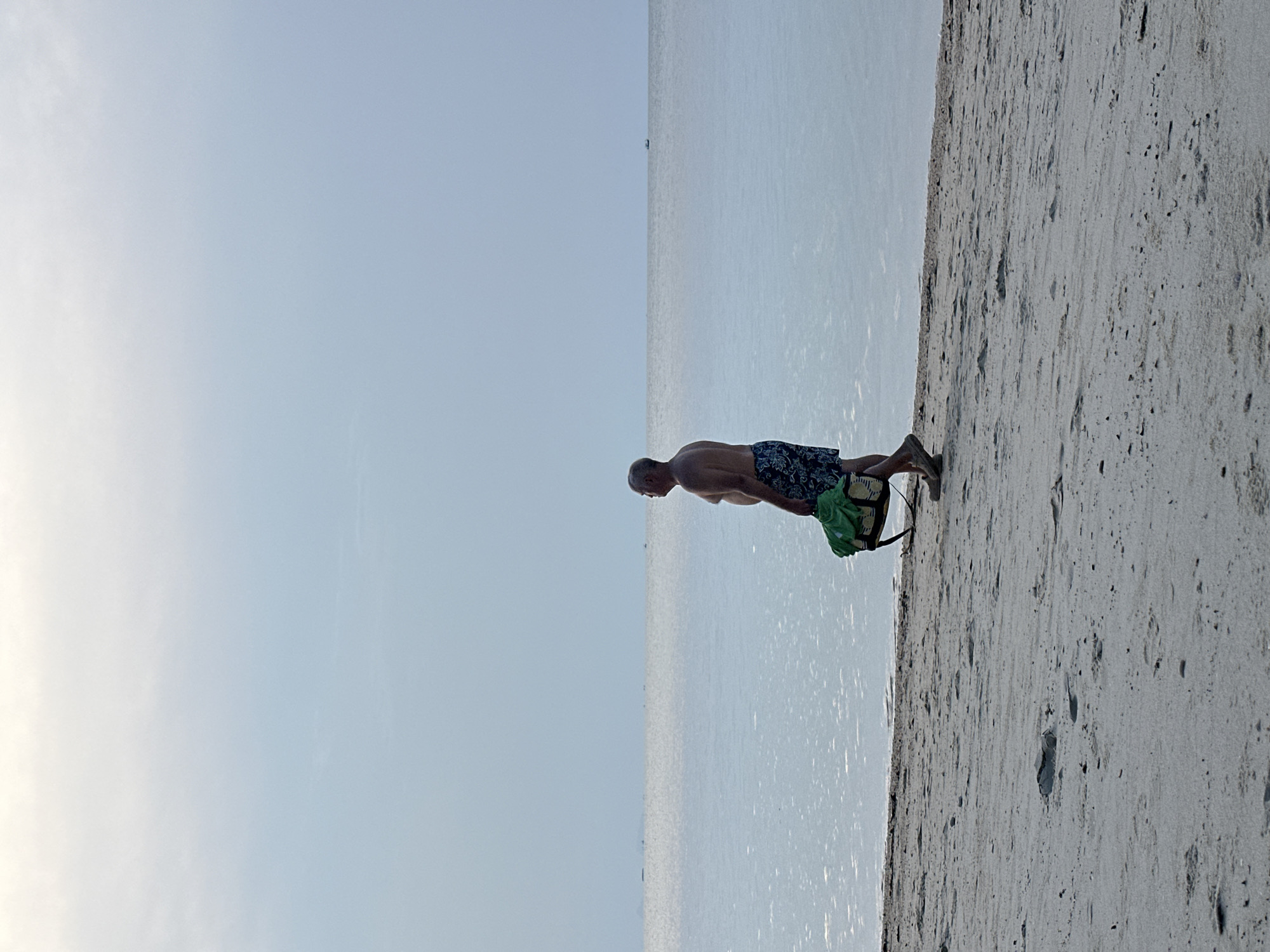 This image captures a serene beach scene, likely in Baan Tai, Thailand, oriented as if rotated 90 degrees clockwise. When viewed upright, the scene depicts a man standing on a sandy beach, facing the sea. The man, shirtless and wearing patterned swim trunks (dark blue, black, and light green/yellow pattern), is bending forward, with his back to the viewer. A green bag or towel rests on the sand near his feet.
The setting is a tranquil beach. The foreground and right side of the image are dominated by light-colored sand, marked with subtle textures and footprints. To the left of the man and extending across the middle of the frame is the calm, light blue sea, reflecting the ambient light and stretching towards a distant horizon. The sky, a soft and clear blue with a lighter hue towards the top left, fills the upper portion of the image. The time of day appears to be bright daytime, possibly late morning or early afternoon, given the gentle illumination and the reflections on the water. The weather is clear and calm. No specific emotions are discernible, and there is no visible text.
