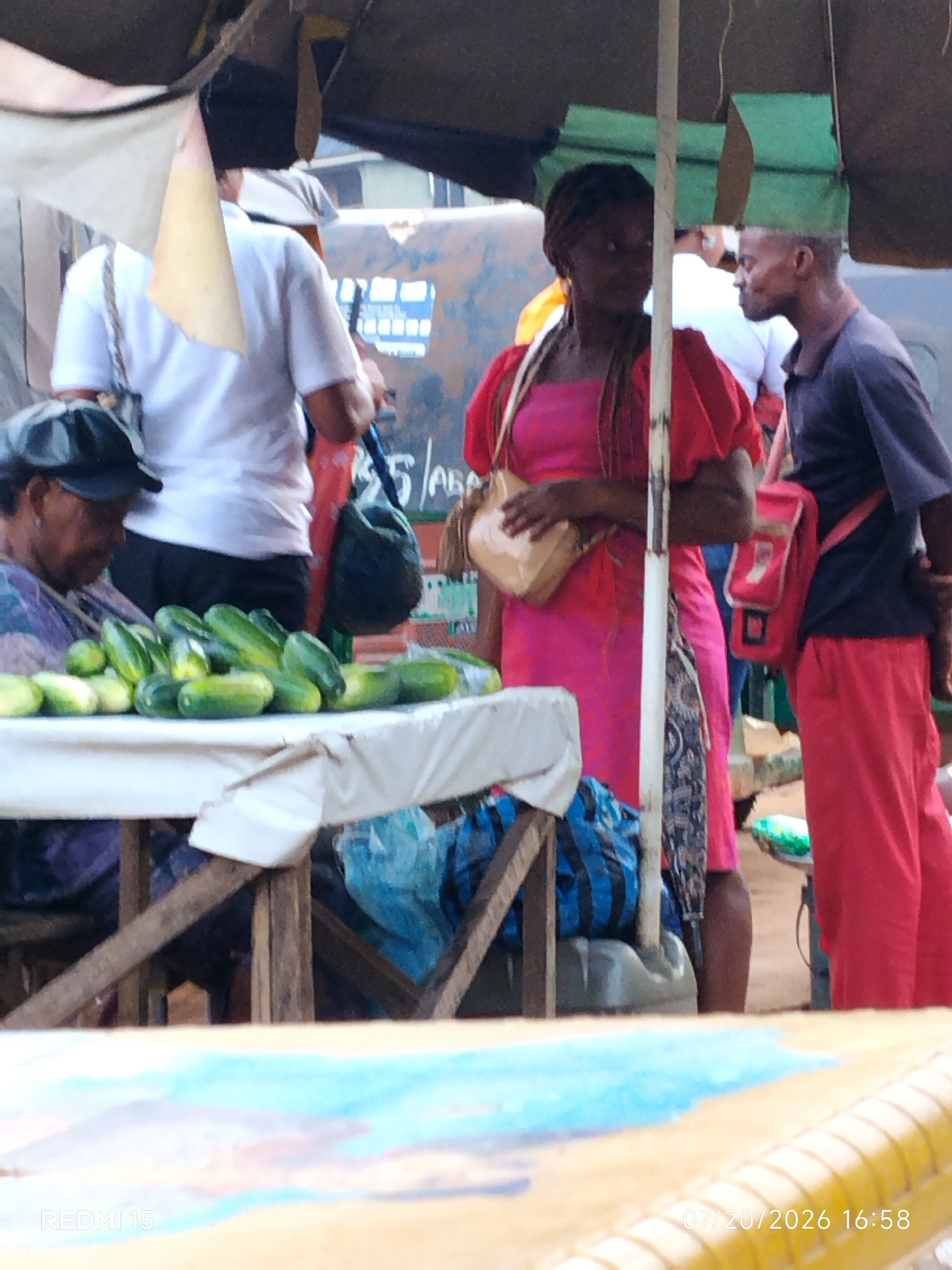 In Umuode, Nigeria, a vendor displays a pile of cucumbers on a table covered with a white cloth. To the left, a woman in a purple top and black cap is partially visible. To her right, a person in a white t-shirt and dark pants stands behind the table. In the center, a woman in a bright pink dress and a man in a dark t-shirt and red pants are positioned. Both are looking in different directions, suggesting they are engaged in interactions related to the market. The environment appears to be an outdoor market with a makeshift canopy overhead, supported by poles. The ground is not clearly visible, but the overall impression is of a bustling marketplace scene.