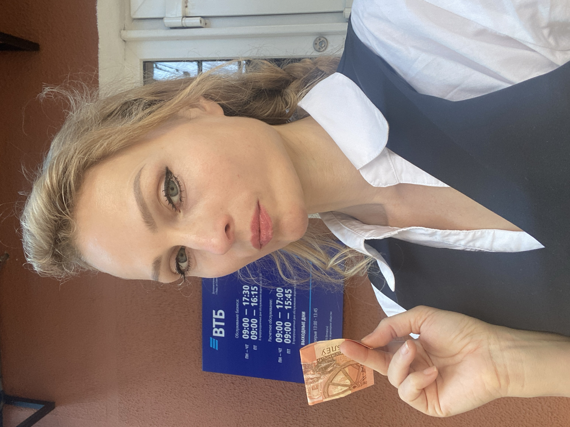 In this medium-shot, waist-up, selfie-style photograph, a fair-skinned woman with blonde hair styled in a messy bun and prominent makeup, including winged eyeliner and pink lipstick, holds a Belarusian 5 Ruble banknote in her right hand. She is positioned in front of a blue sign with white text, which appears to be outside a VTB bank branch, identified by the text "ВТБ" on the sign. The sign details the bank's operating hours for business and settlement services, indicating it is located in Lida, Belarus, as suggested by the text within the cropped image which reads "в г. Лида, ул. Гоголя" (in Lida city, Gogol St.). The woman is wearing a white collared shirt and a dark V-neck vest over it. The background also shows a portion of a building facade and a white door or window frame. The overall lighting suggests an outdoor setting during daytime.