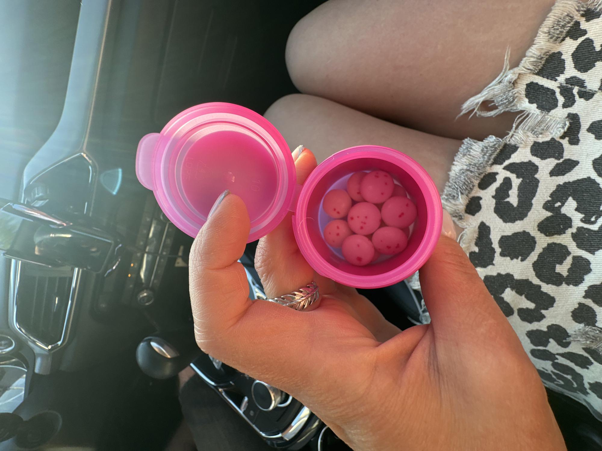 A person is sitting in a car, holding a pink container with pink round objects in it. The container lid is open and next to the container. The person is wearing a silver ring with a leaf design. The person is wearing a leopard print skirt. The car interior is visible in the background, including the dashboard, steering wheel, and gear shift. The car is parked in an unknown location. There is no evidence to indicate the location is the city of Chaikovsky, Russia.