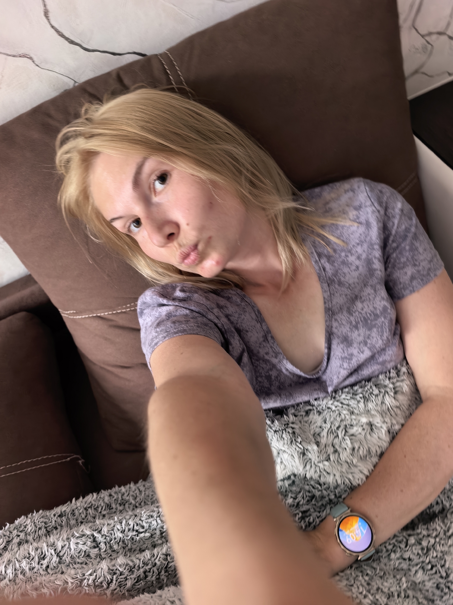 A fair-skinned blonde woman is visible in a close-up selfie taken indoors. She is in repose, seemingly lying on a brown plush surface with a gray and white fluffy material covering part of her body. She is wearing a gray V-neck T-shirt. On her left wrist, a smartwatch displays the time 16:09. The background includes a wall with a marble-like pattern and a dark-colored surface. The location in the city of Sterlitamak, Russia is unknown.