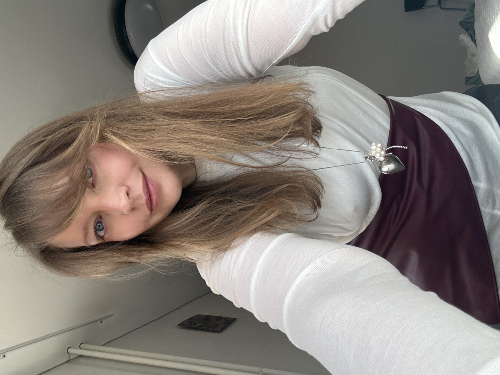 The image depicts a fair-skinned woman, approximately 25-35 years old, with long light brown hair styled with bangs, blue eyes, and light pink lips, captured in a close-up selfie from a low angle. She exhibits a slight smile and looks directly into the camera. She is wearing a white, long-sleeved top, under which a dark burgundy or maroon garment, possibly a wide belt or part of a skirt, cinches her waist. A silver-colored necklace with two pendants, one a polished heart and the other a cluster of white pearls with a silver accent, rests on her chest.
The scene is indoors, in what appears to be a residential setting within Izhevsk, Russia. The background features light-colored walls and ceiling. Visible on the left wall are two thin, light-colored vertical cylindrical objects, possibly pipes, attached near the ceiling, and a dark rectangular object, likely a framed picture or wall hanging. In the upper right background, a circular light fixture or ceiling fan is partially visible on the ceiling. Towards the bottom right, part of a light-colored bed with white bedding and a dark chair are visible, suggesting a bedroom environment. The lighting is soft and even, likely a mix of natural and artificial light.