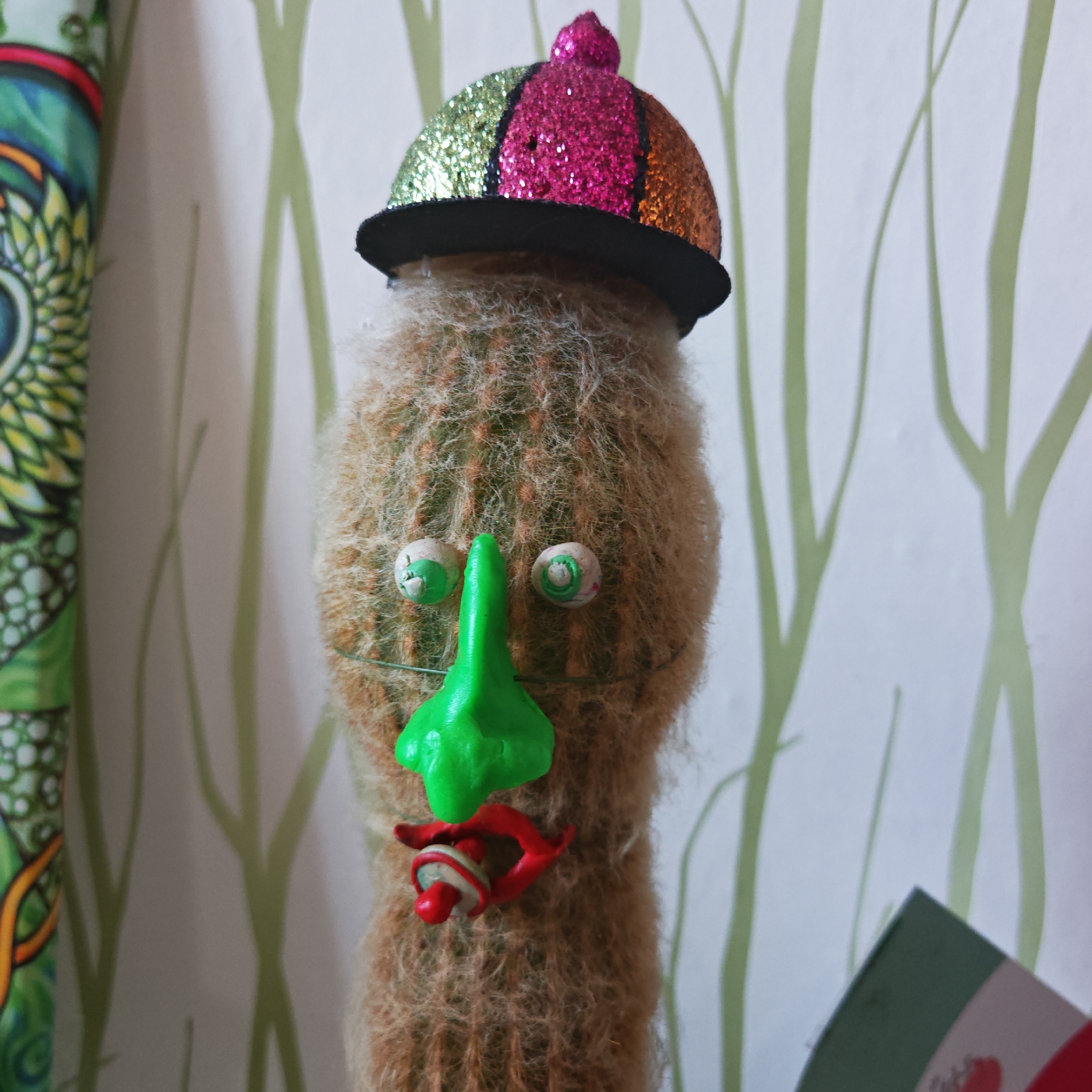 A close-up shows a cactus decorated to look like a comical face with a multicolored, glittery hat on top. The hat is divided into sections of green, pink, and gold glitter. The cactus "face" is formed by crude craft additions: large, cartoonish eyes made of white clay with green swirls, a prominent green nose, and a twisted red mouth. The background is a blurry, white wall with light green, leafy patterns. A portion of a patterned pole or fabric is visible to the left, and the edge of what appears to be a flag (possibly Mexican) is visible to the right. The photograph was probably taken indoors. The location is not discernible.