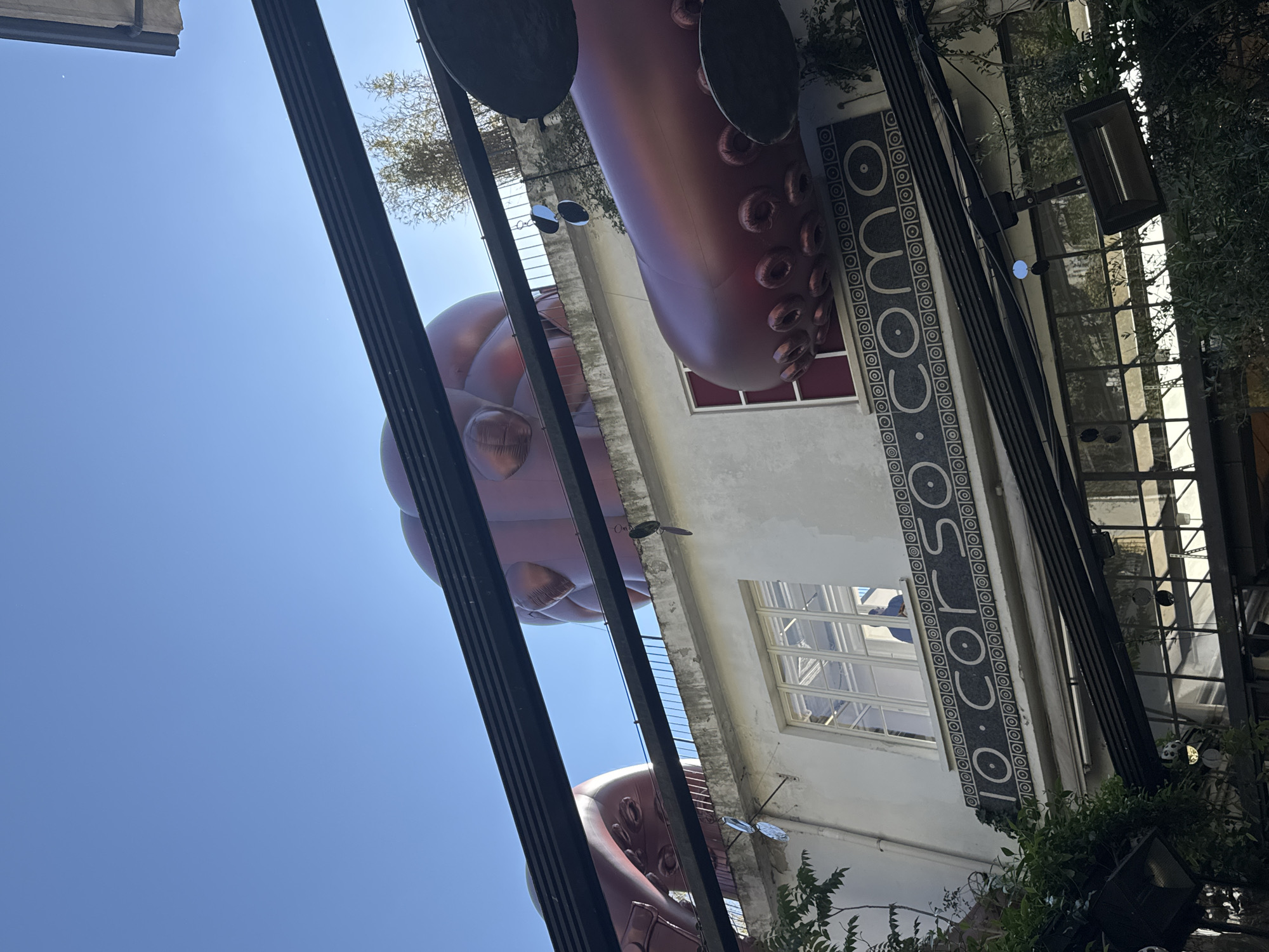 The image captures a street scene in Milan, Italy, specifically at "10 Corso Como." A large, inflated sculpture of an octopus is prominently displayed above, partially obscuring the sky. The octopus is reddish-brown with visible suction cups on its tentacles and reflective circular elements. The building is light-colored with windows. There's a sign vertically oriented on the wall with the inscription "10 CORSO COMO." Metal framework structures are visible in the foreground, and foliage from plants is also present. The sky is clear and blue.