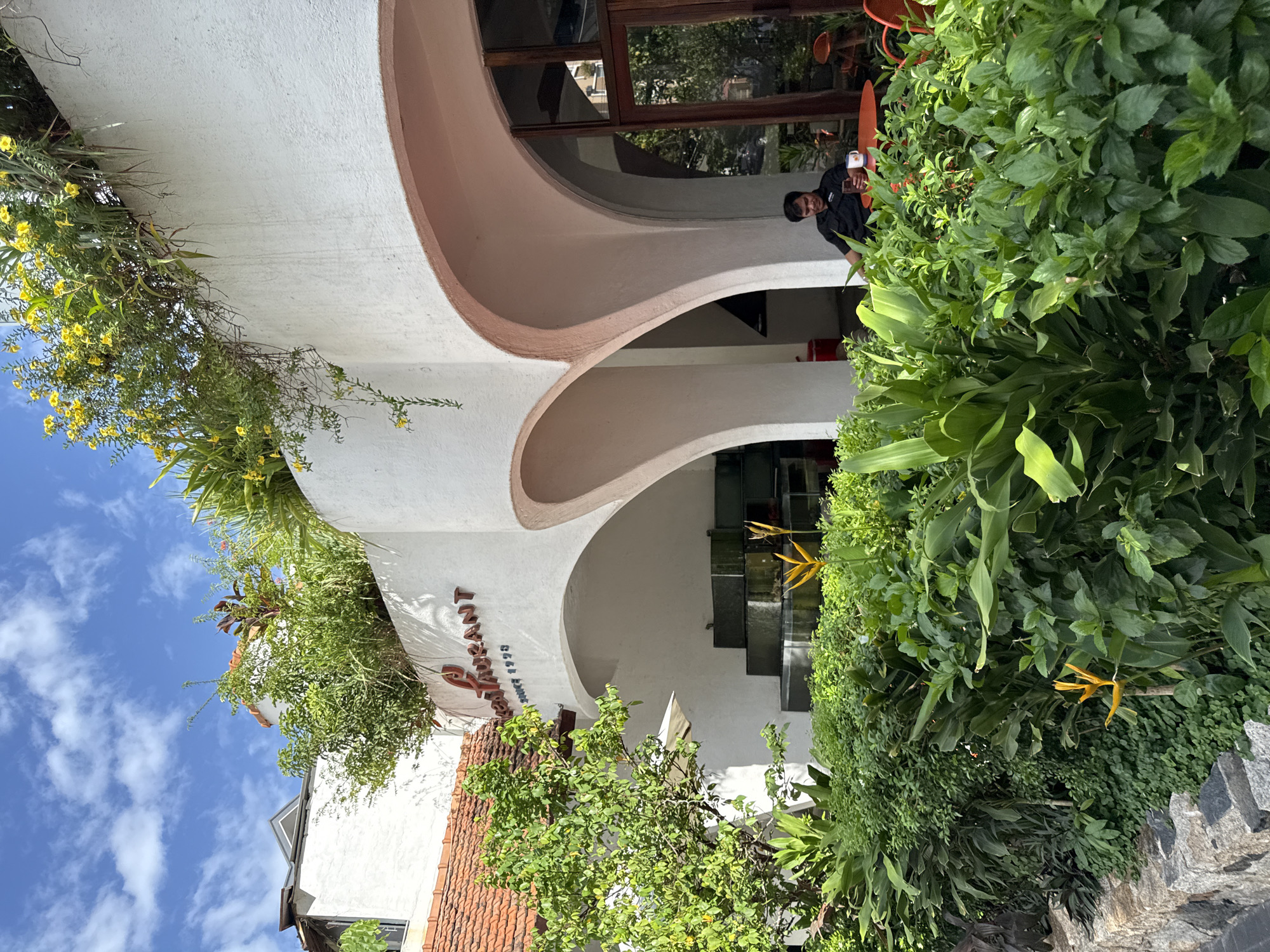 The image shows a street view in Nha Trang, Vietnam, featuring a restaurant with a distinctive curved architectural design and lush greenery. The restaurant's facade is painted white and adorned with brown lettering that reads "Restaurant Since 1999". Above the building, a green terrace with blooming yellow flowers adds to the vibrant scenery. In the foreground, a variety of plants, including large tropical leaves and small ground cover, are visible. A stone wall borders the walkway in the lower left corner. Inside the covered outdoor seating area, a man wearing a black shirt is seated at an orange table, holding a smartphone and a mug. The sky above is blue with scattered white clouds. The overall scene suggests a warm, sunny day in a tropical urban setting.