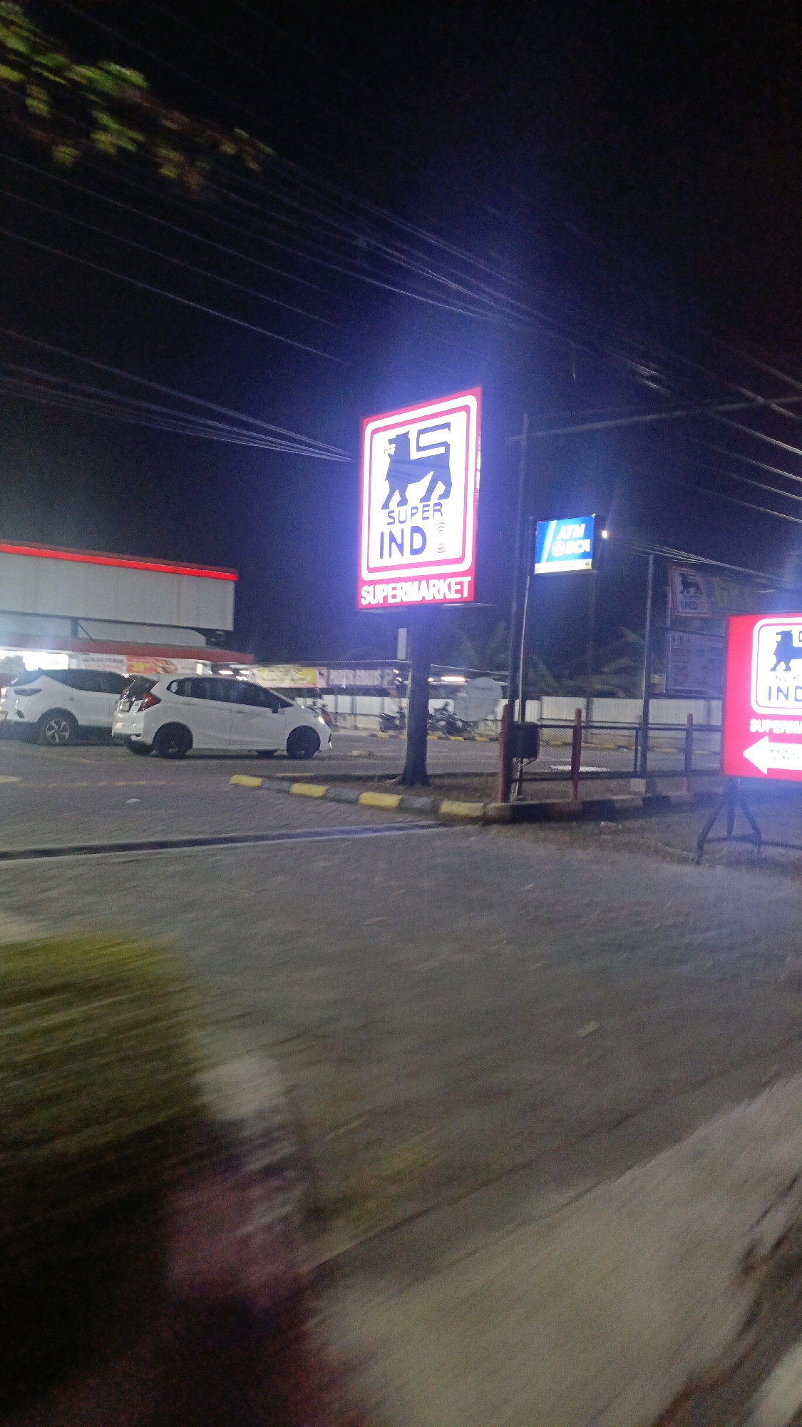 The image depicts a nighttime view of a "SUPER INDOMARET" supermarket located on a street in Siswodipuran, Indonesia. A prominent illuminated sign for "SUPER INDO" with a lion logo is visible. To the right of this sign, another illuminated sign for "ATM BCA" is present. Several white cars are parked in the lot in front of the supermarket. The street and parking lot are paved, and there are yellow and black speed bumps. In the background, the supermarket building appears to be a single-story structure with a red-trimmed roof. The scene is illuminated by the artificial lights of the signs and the building. The image appears to have been taken from a moving vehicle due to motion blur in the foreground.