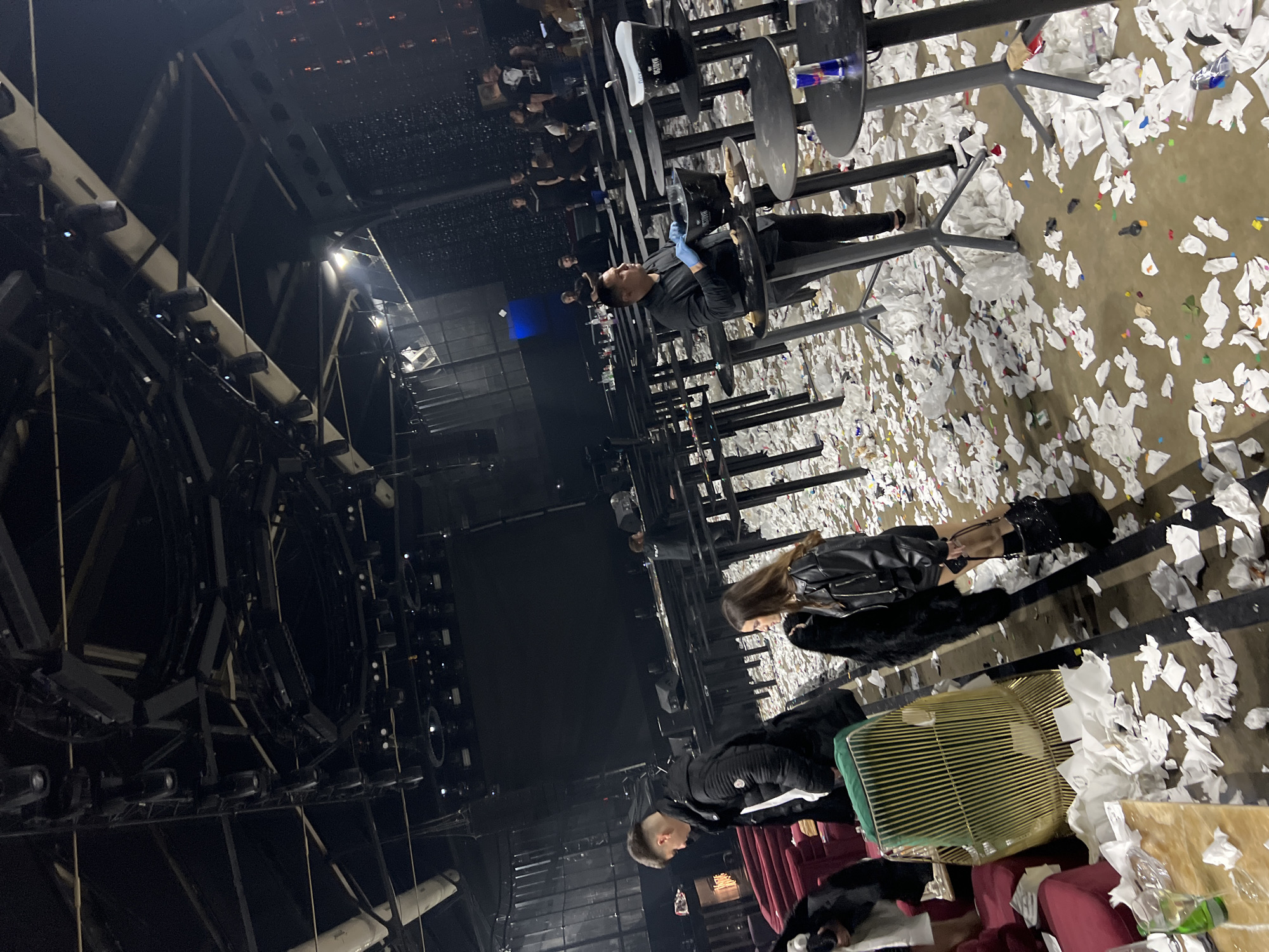 The image depicts the interior of what appears to be a nightclub or event space, possibly in Burgas, Bulgaria. The floor is covered in a heavy layer of shredded paper, creating a confetti-like effect. Several people are present, some standing or sitting at tables, others interacting with the environment. The tables are black with circular tops, and some have drink cans or other items on them. A person wearing blue gloves is visible near a table. A woman in a black leather jacket is standing. Near the lower portion of the image, a person stands near a red curtain and furniture, which includes an ornate birdcage, suggesting a theatrical element to the space. The ceiling is high and complex, with visible lighting rigs and support structures.