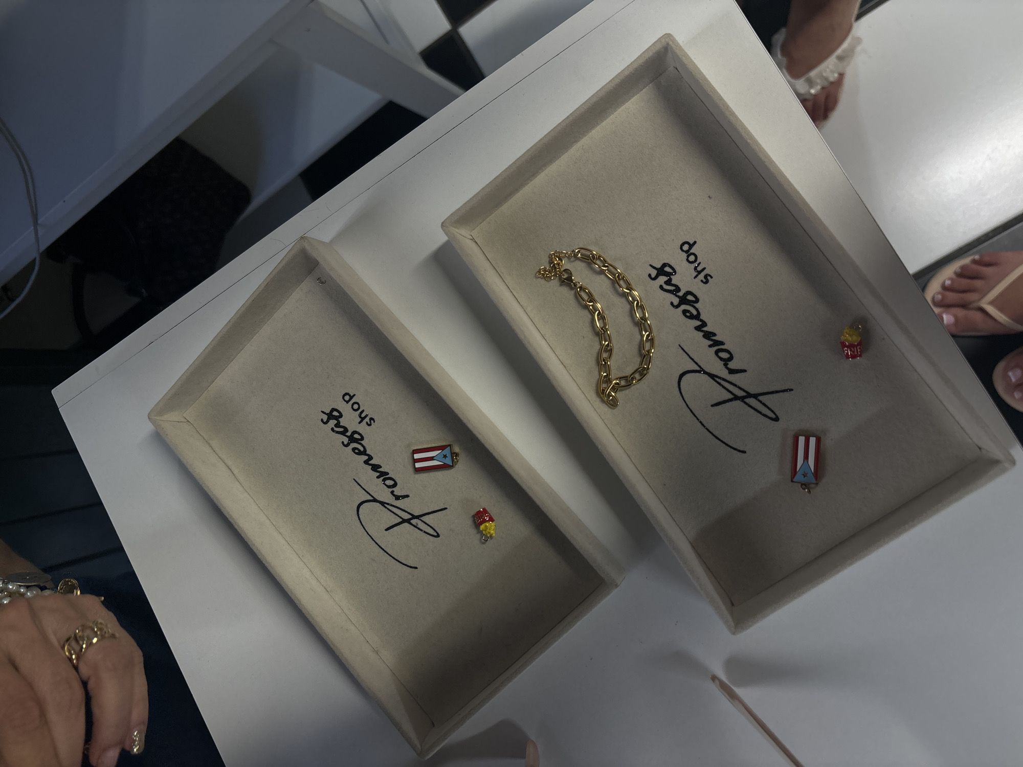 In San Juan, Puerto Rico, on a white surface, two tan fabric-lined trays are displayed. The upper tray contains two small charms: one is a miniature Puerto Rican flag, and the other is a red and yellow object resembling a hot dog or french fries. A logo that reads "Promesas shop" is printed on the tray. The lower tray features a gold chain bracelet. It also has a logo similar to the upper tray and a miniature Puerto Rican flag charm. A person's hands are visible in the upper left corner, wearing jewelry. Another person's feet are visible in the bottom right corner, wearing flip-flops. The background includes black and white checkered flooring and a black object partially visible in the upper right.