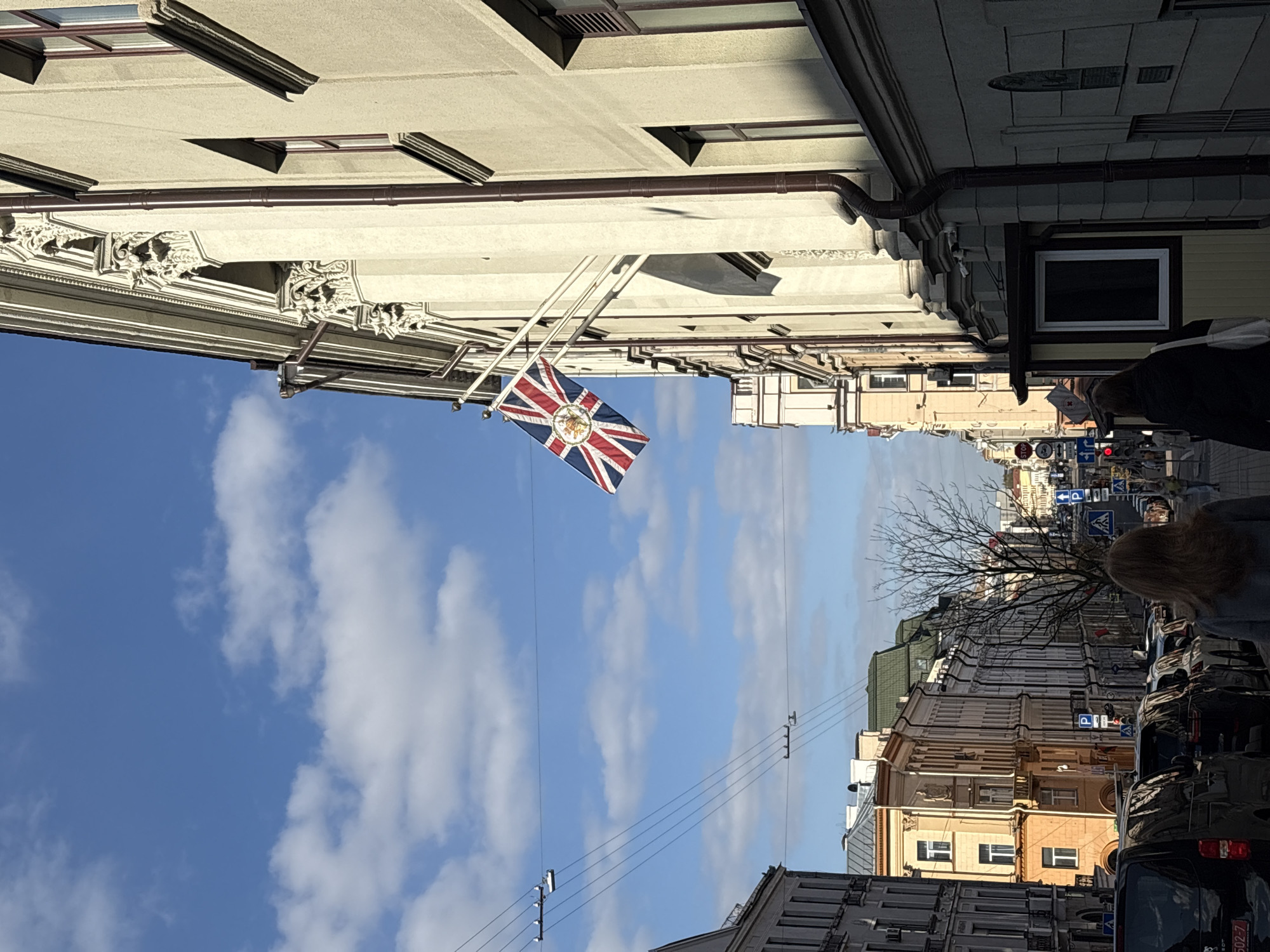 This image captures a vibrant street scene in a European city, with a prominent British flag hanging from an ornate building. The perspective is looking down a city street, flanked by multi-story buildings under a clear, bright blue sky dotted with white clouds, suggesting a sunny day.
On the left side of the street, a grand, light-colored building features intricate architectural details, including decorative corbels and moldings under its roofline. A large flag of the United Kingdom (Union Jack) with the Royal Arms of the United Kingdom centered on it is prominently displayed, hanging from this building on a pole. This specific flag design, typically used by British diplomatic missions, strongly indicates the presence of the British Embassy or Consulate.
The street itself appears paved, with numerous cars parked along the right side. People are visible walking along the sidewalks further down the street, indicating a normal day's activity. A bare-branched tree stands on the right, suggesting either late autumn or winter. The buildings on the right side of the street vary in style, some appearing older with classic architecture, others perhaps renovated or more modern.
Overhead, electrical or telephone lines crisscross the street. Several street signs are discernible, including blue 'P' signs indicating parking and a red 'STOP' sign. The visible text on the 'STOP' sign is in Cyrillic, further supporting the location in an Eastern European country. This, combined with the provided context, places the scene specifically in Minsk, Belarus, likely depicting the British Embassy. The overall impression is one of a bustling city street on a clear, pleasant day.