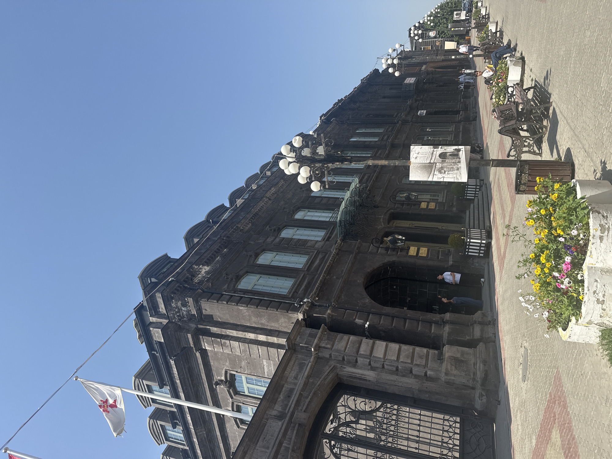 The image shows a view of a street in Gyumri, Armenia, on a sunny day. The architecture is predominantly dark stone, with ornate details and mansard roofs. A white flag with a red cross is flying from a flagpole in front of a building on the left. The street is paved with red and white bricks and features rows of white lampposts with multiple lights. Benches are placed along the sidewalk, some occupied by people. Flower beds with colorful flowers are also visible. On the right side of the street, there are more buildings with similar architectural styles, and a line of trees extends into the distance. Some people are walking on the sidewalk, while others are sitting on the benches. A trash can is positioned near some of the benches. The sky is clear and blue.