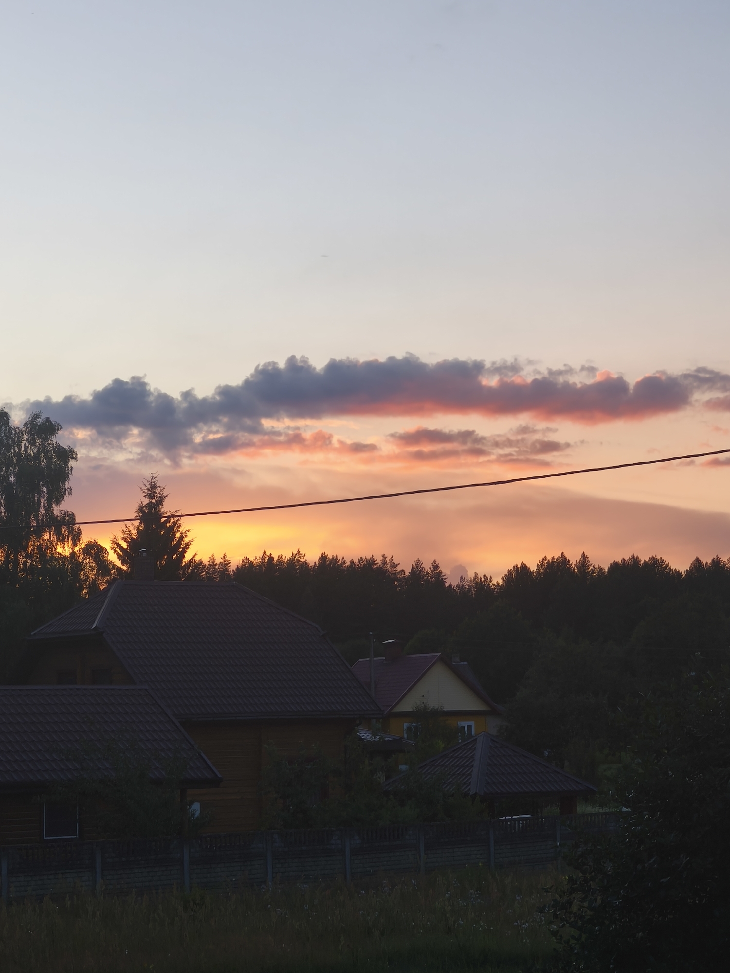 The image depicts a rural landscape at dusk in Poreccha, Belarus. A warm sunset bathes the sky in hues of orange and pink, with scattered clouds. In the foreground, two wooden houses with dark tiled roofs are visible, partially obscured by trees. A fence runs along the property line, separating the houses from a grassy area. A dense line of dark green trees forms the horizon behind the houses. The overall impression is one of a peaceful, tranquil evening in a rural setting.
