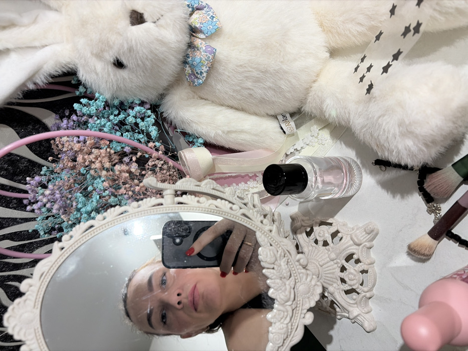 A woman's reflection appears in an ornate, oval mirror surrounded by various objects. A plush white rabbit with a floral bow lies near a bouquet of colorful dried flowers. Next to the mirror, a perfume bottle rests near a makeup brush. A pink headband, ribbon, and lace details are visible. The woman holds a smartphone, capturing the scene in the mirror, her red nail polish catching the light. The backdrop is a zebra-striped pattern.