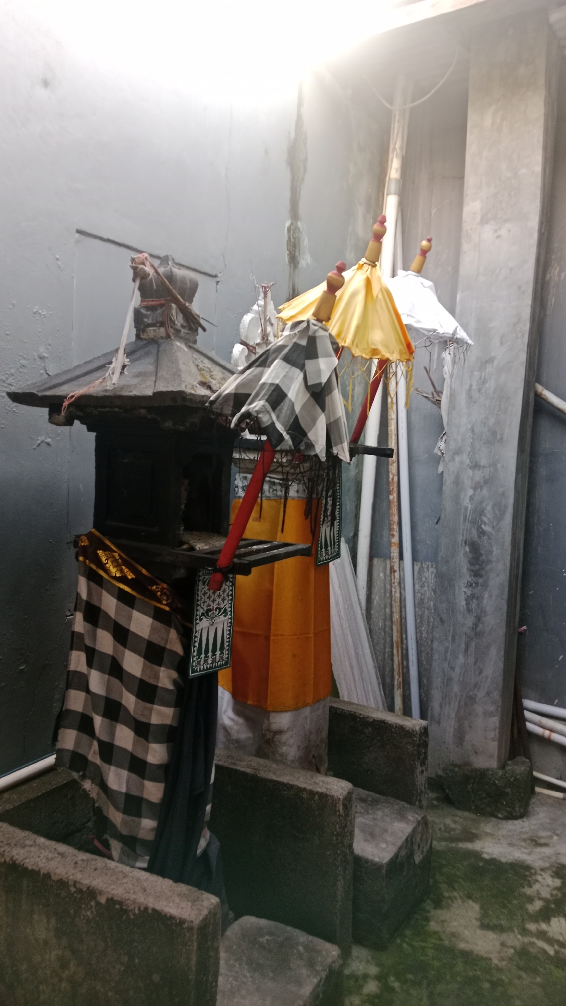 The image shows a traditional Balinese offering structure adorned with checkered and yellow fabrics, along with ceremonial umbrellas. The structure is placed outdoors, possibly in a courtyard or alleyway, with concrete blocks and walls forming the surroundings. The scene appears to be in Denpasar, Indonesia, suggested by the cultural elements and architectural style. The lighting indicates it's daytime, with strong sunlight casting a glare. No people are visible in the frame.