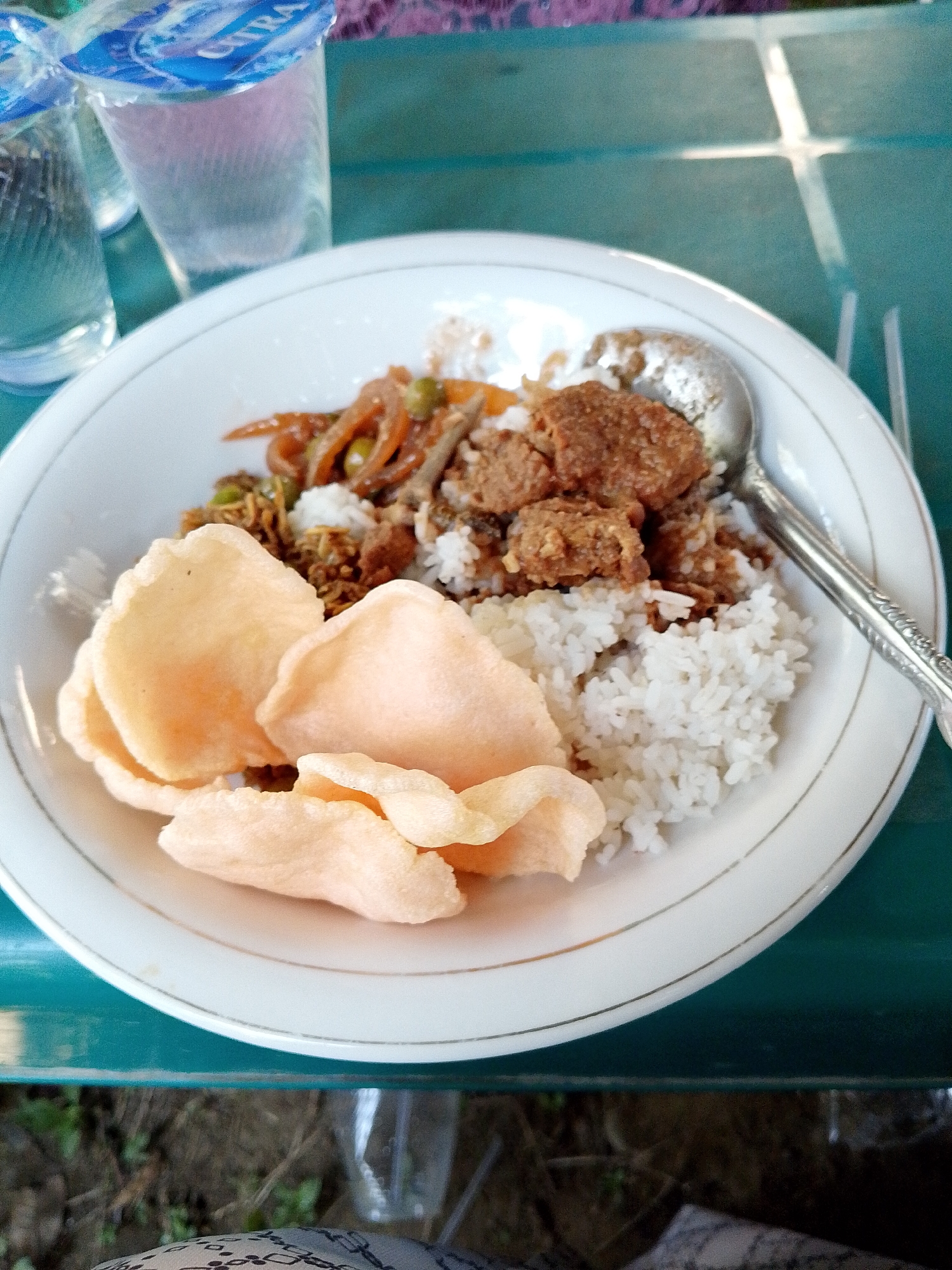 A close-up, eye-level shot shows a white plate of food on a teal-colored table in Meunasah Kulam, Indonesia. The plate contains a generous portion of white rice, topped with several pieces of fried meat and a side of stir-fried vegetables that include sliced carrots and green peas. Three light-pink crackers are arranged in front of the rice. A silver spoon rests on the right side of the plate, partially submerged in the rice. In the background, two glasses of water with blue lids are visible, as well as a hint of a patterned cloth. The table surface has a subtle, reflective sheen. The overall lighting suggests an outdoor setting with natural light.