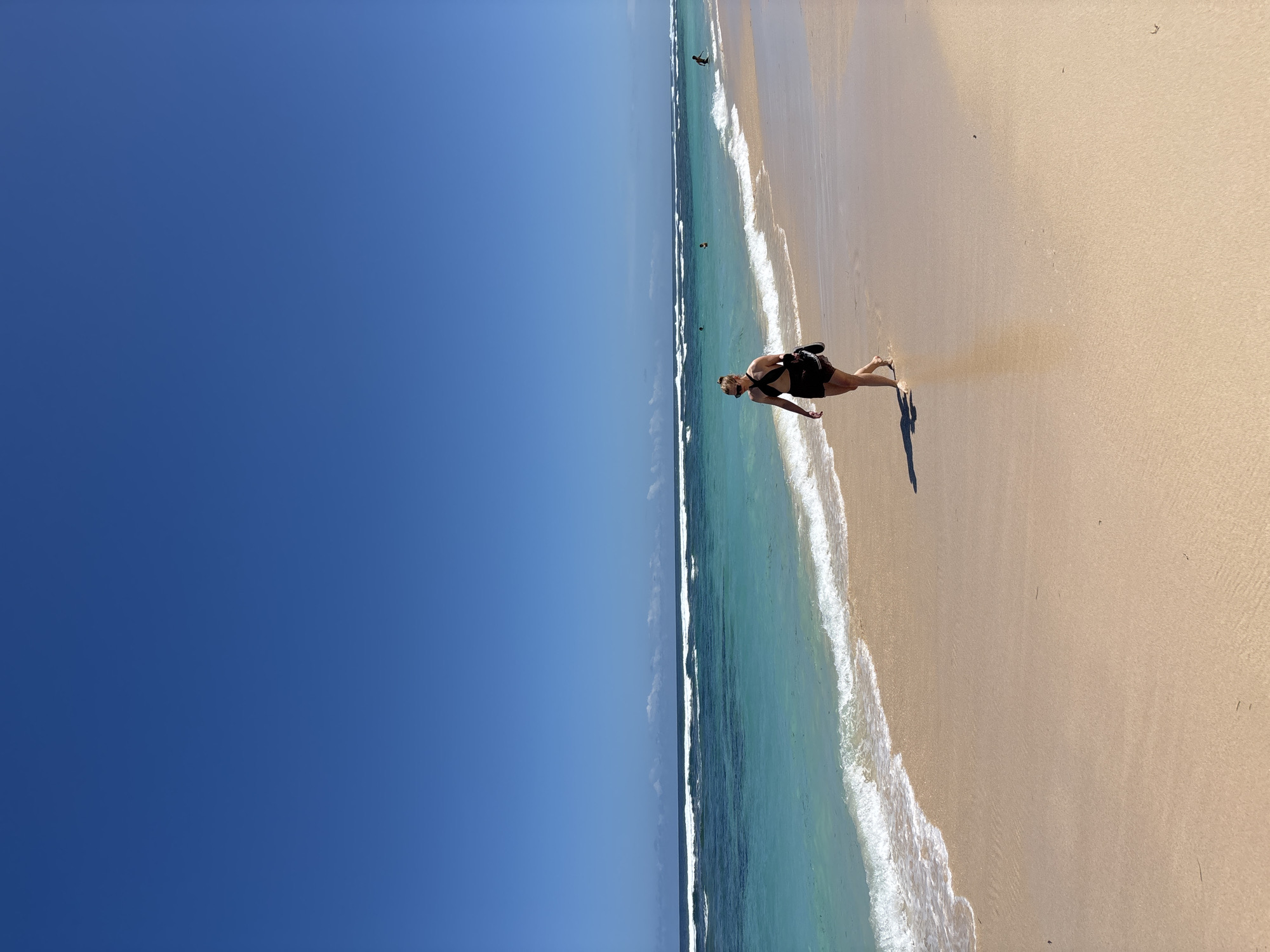 The image shows a beach scene, likely in Kuta Selatan, Indonesia, based on the landscape characteristics. A woman in dark swimwear and a dark shoulder bag is standing on the wet sand near the shoreline. The ocean is turquoise, with white waves breaking. The sand is tan-colored. The sky is a clear, deep blue. There are other people in the water further down the beach. The image is oriented vertically.