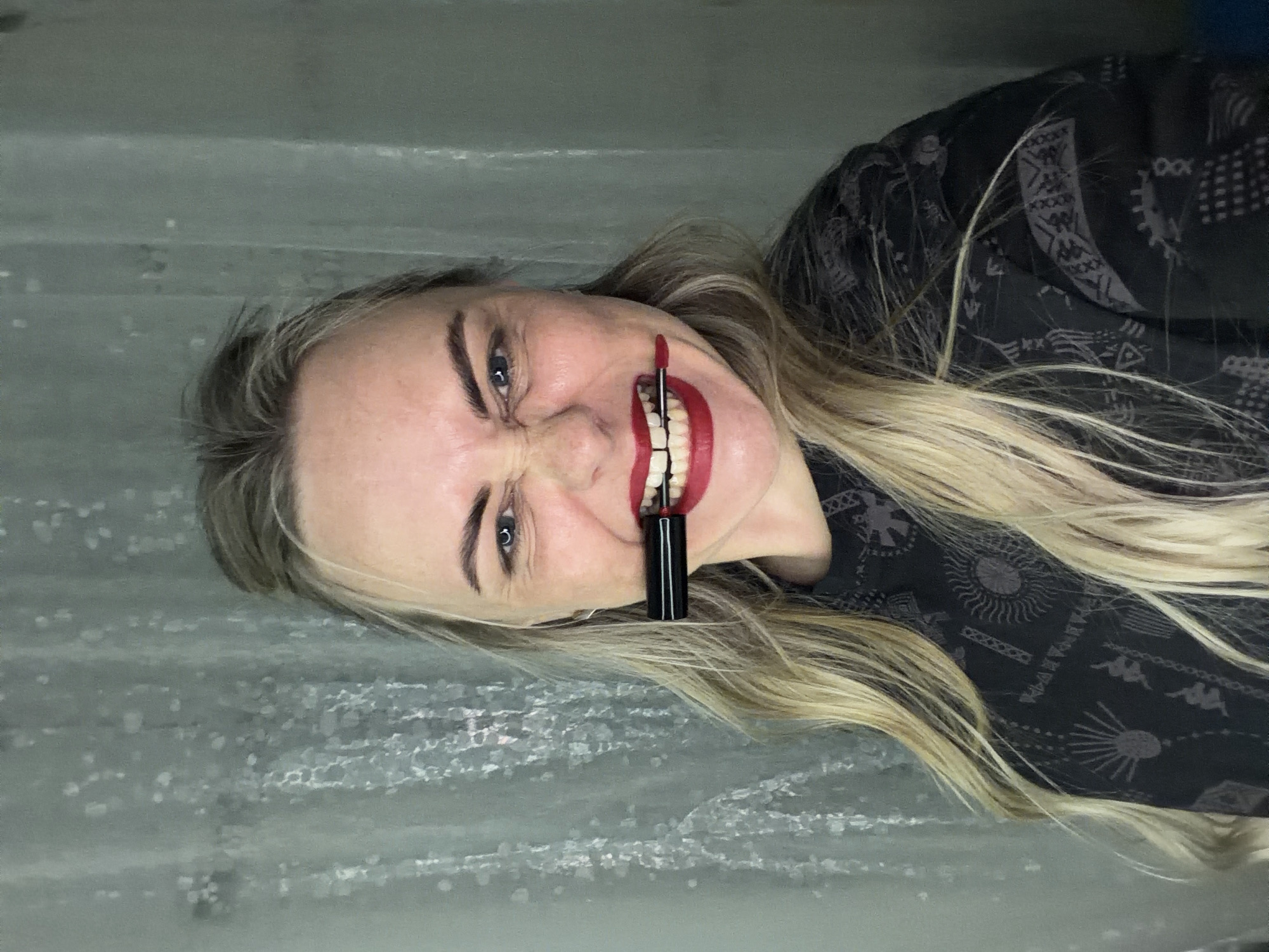 A woman lies on a gray surface, possibly concrete. She has blonde hair and is wearing a black top with a repeating gray geometric pattern. She is holding a tube of red lipstick in her mouth, painting red lipstick across her teeth and lips with a smile. The background suggests an outdoor setting, though there are no specific details to discern a precise location in Votkinsk, Russia, if applicable.